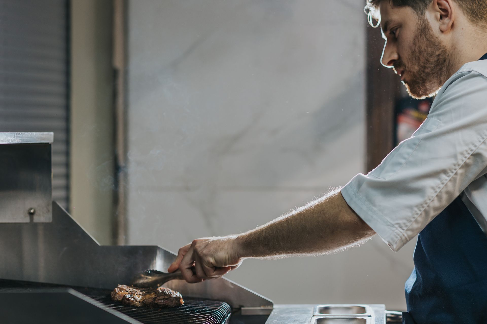 Chef grilling meat on a stainless steel grill. He is wearing a white coat and blue apron.