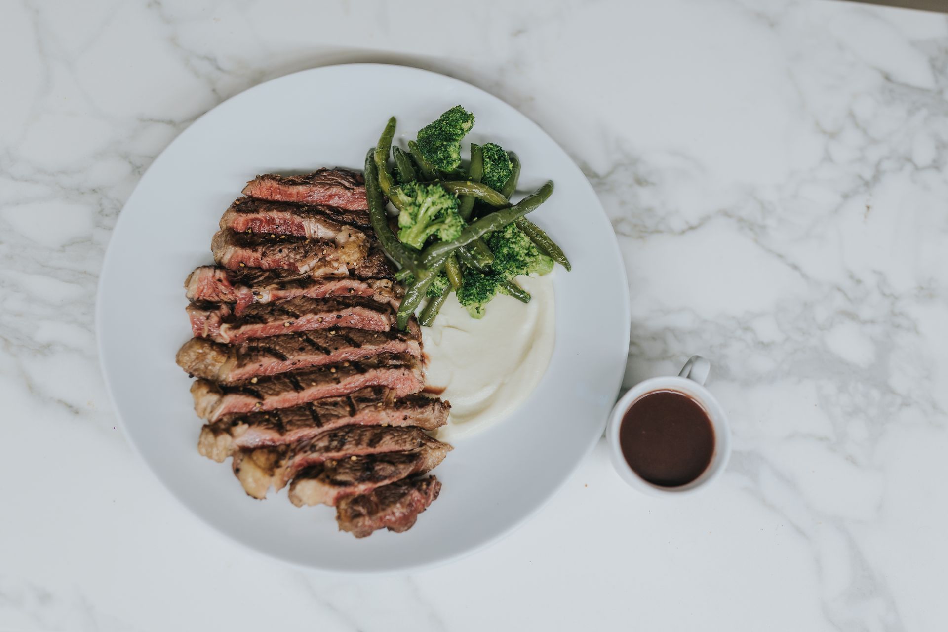 Sliced steak with mashed potatoes, broccoli, green beans, and sauce on a white plate.