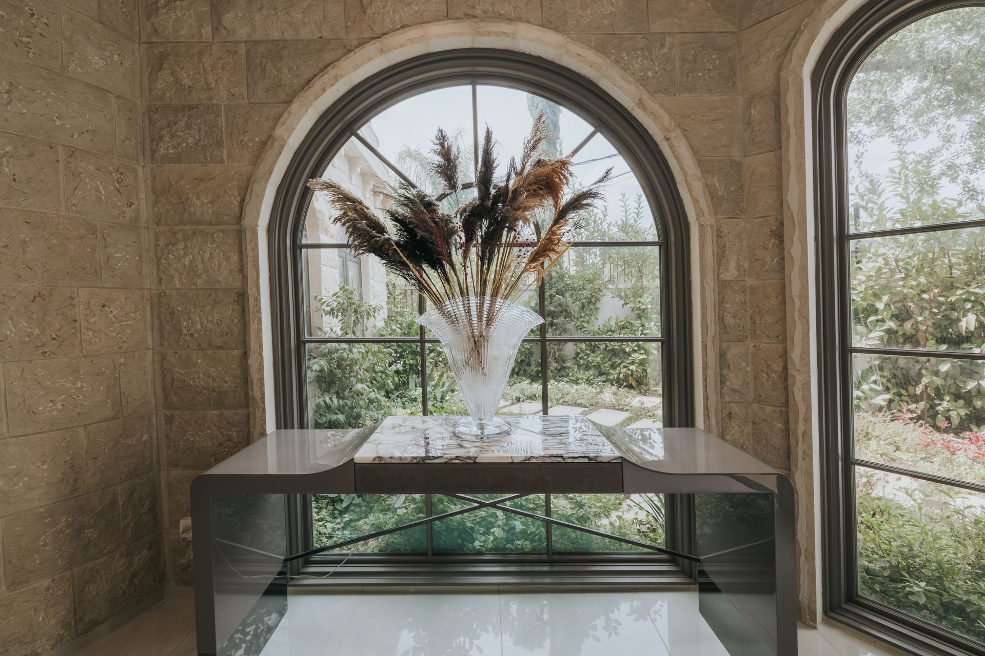 A console table with a vase of dried flowers in front of an arched window.