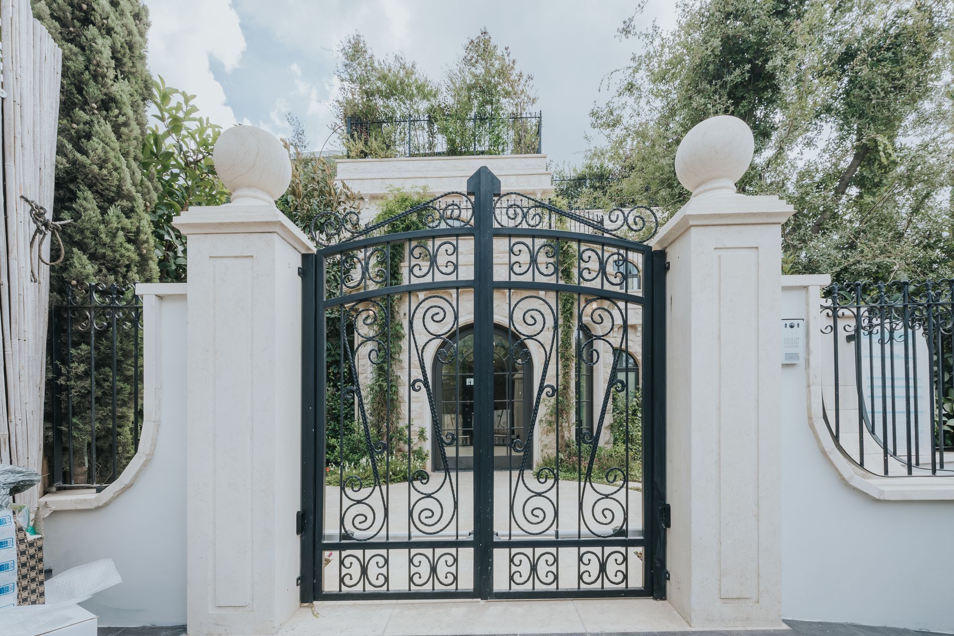 Black wrought-iron gate within white stone walls, topped with spherical finials. Lush greenery visible behind.