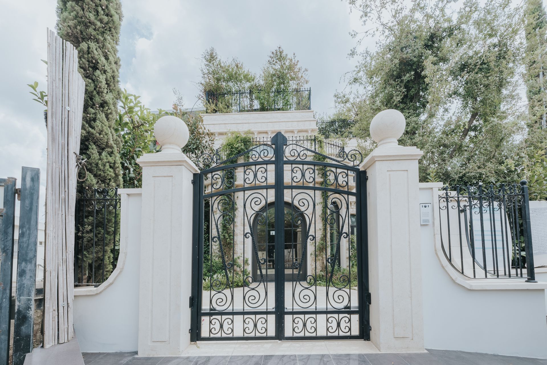 Black wrought-iron gate with decorative swirls, set between white pillars with stone spheres, leading to a building.