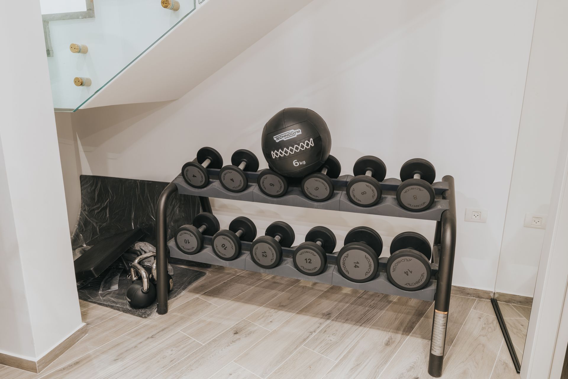 Dumbbells and medicine ball on a rack, indoors near a staircase. The rack is black.