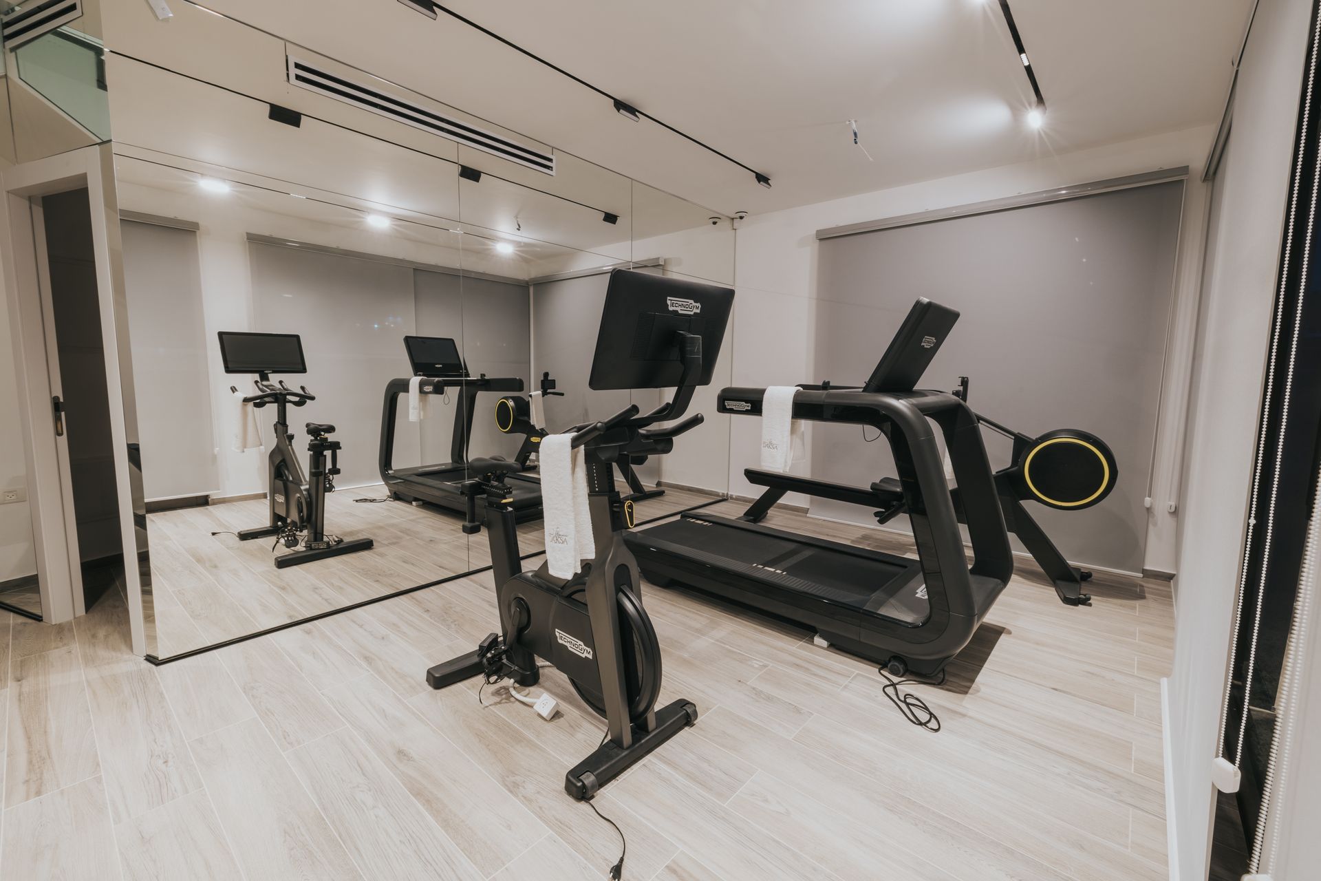 Gym with treadmills, stationary bikes, and a large mirror. Light wood floors, neutral walls, and a window with blinds.