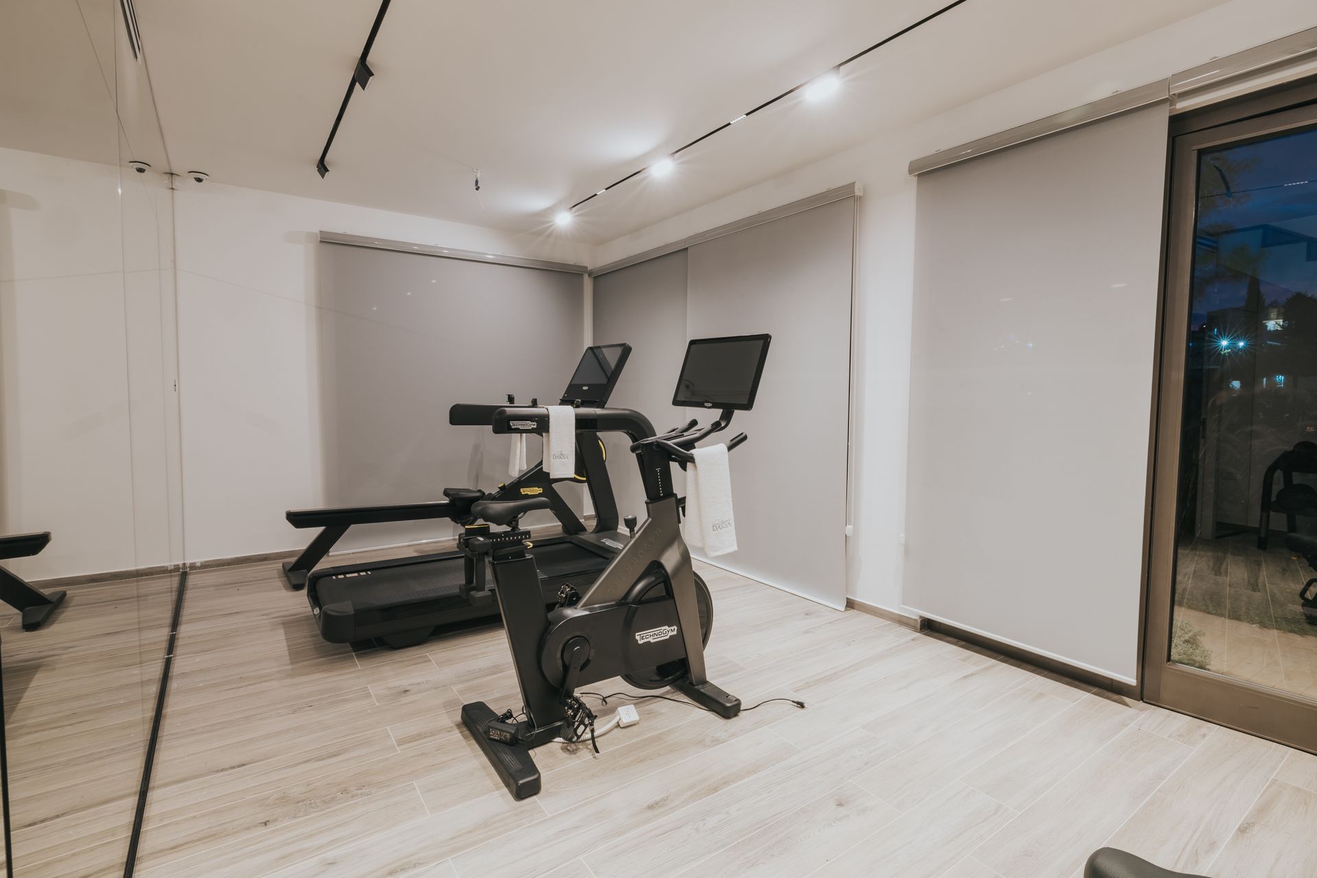 Gym with exercise bike, treadmill, and gray window shades. Wooden floor, neutral walls, and track lighting.