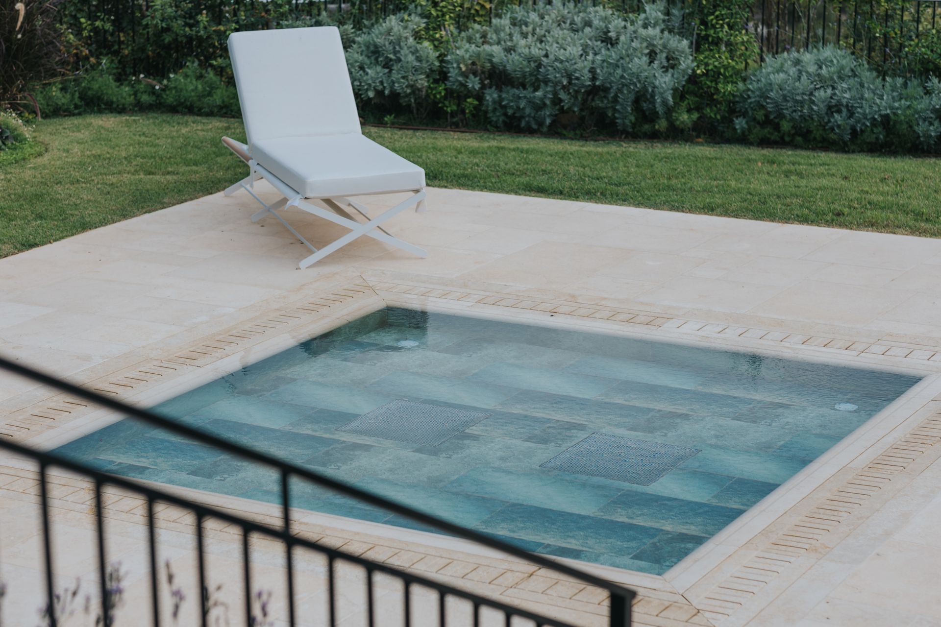 Square pool with turquoise water, beige stone surround, and white lounge chair on grass.