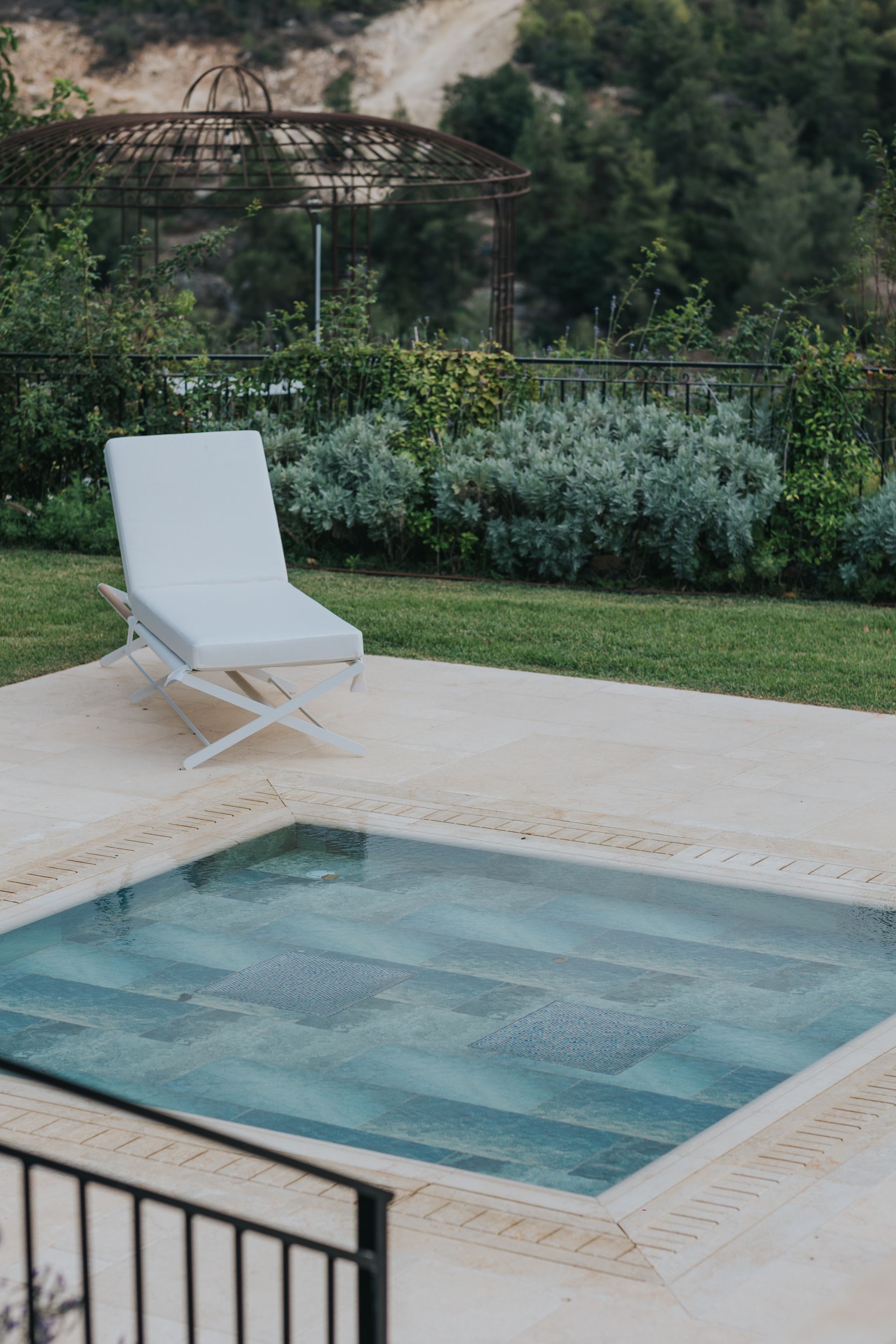 Poolside scene with white lounge chair next to a rectangular pool, surrounded by greenery.
