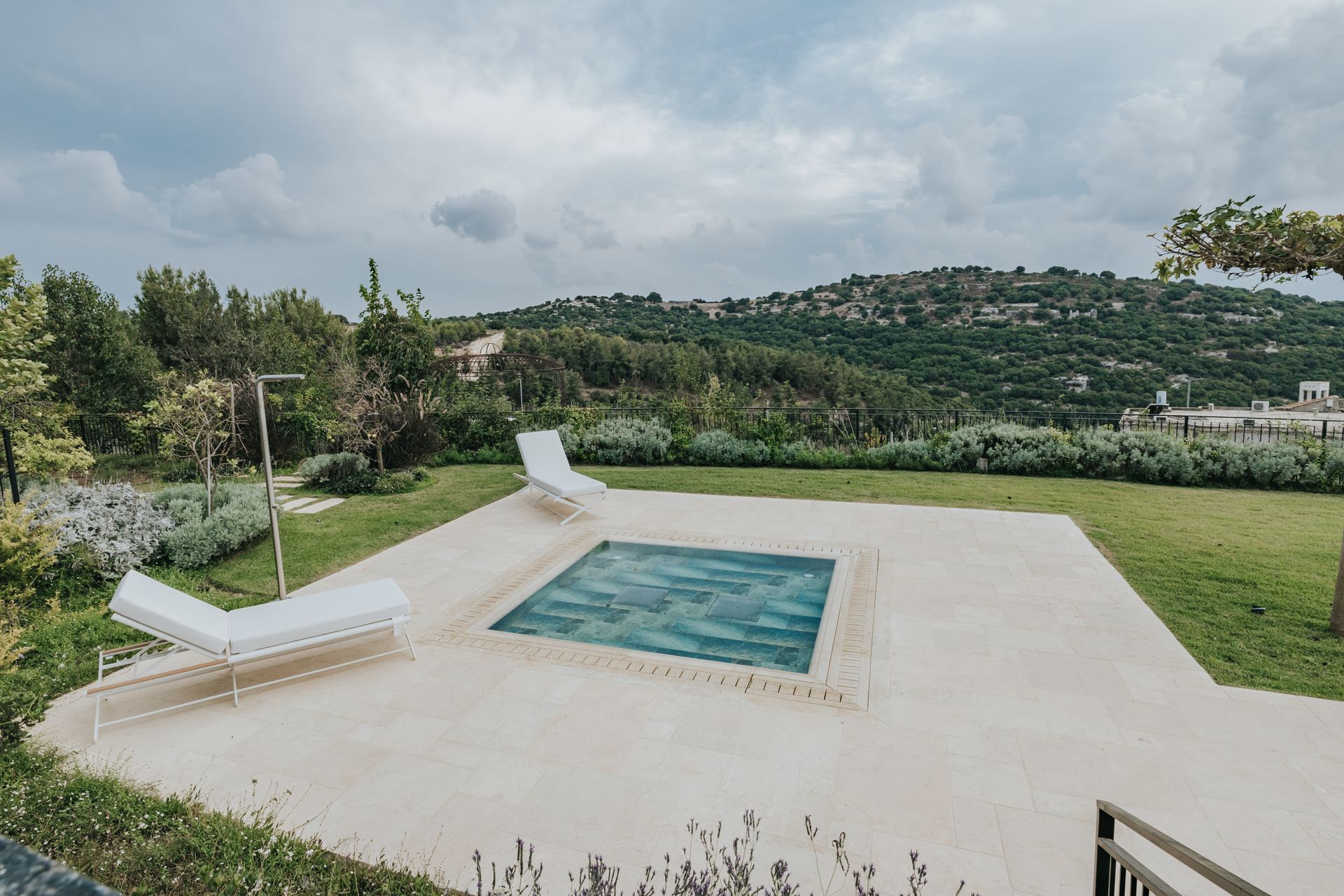 Pool with blue tile surrounded by a concrete patio and two white lounge chairs, with a green lawn and hills in the background