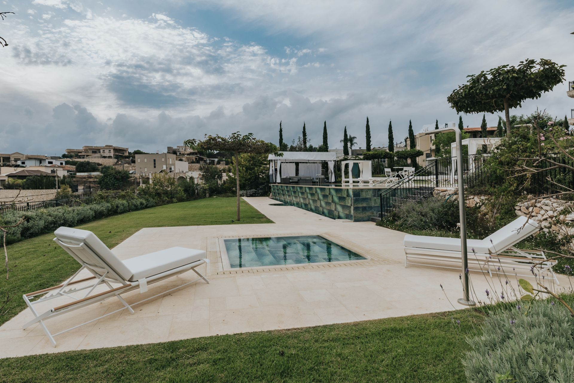 Lounge chairs beside a small pool in a yard, overlooking a distant village and cloudy sky.
