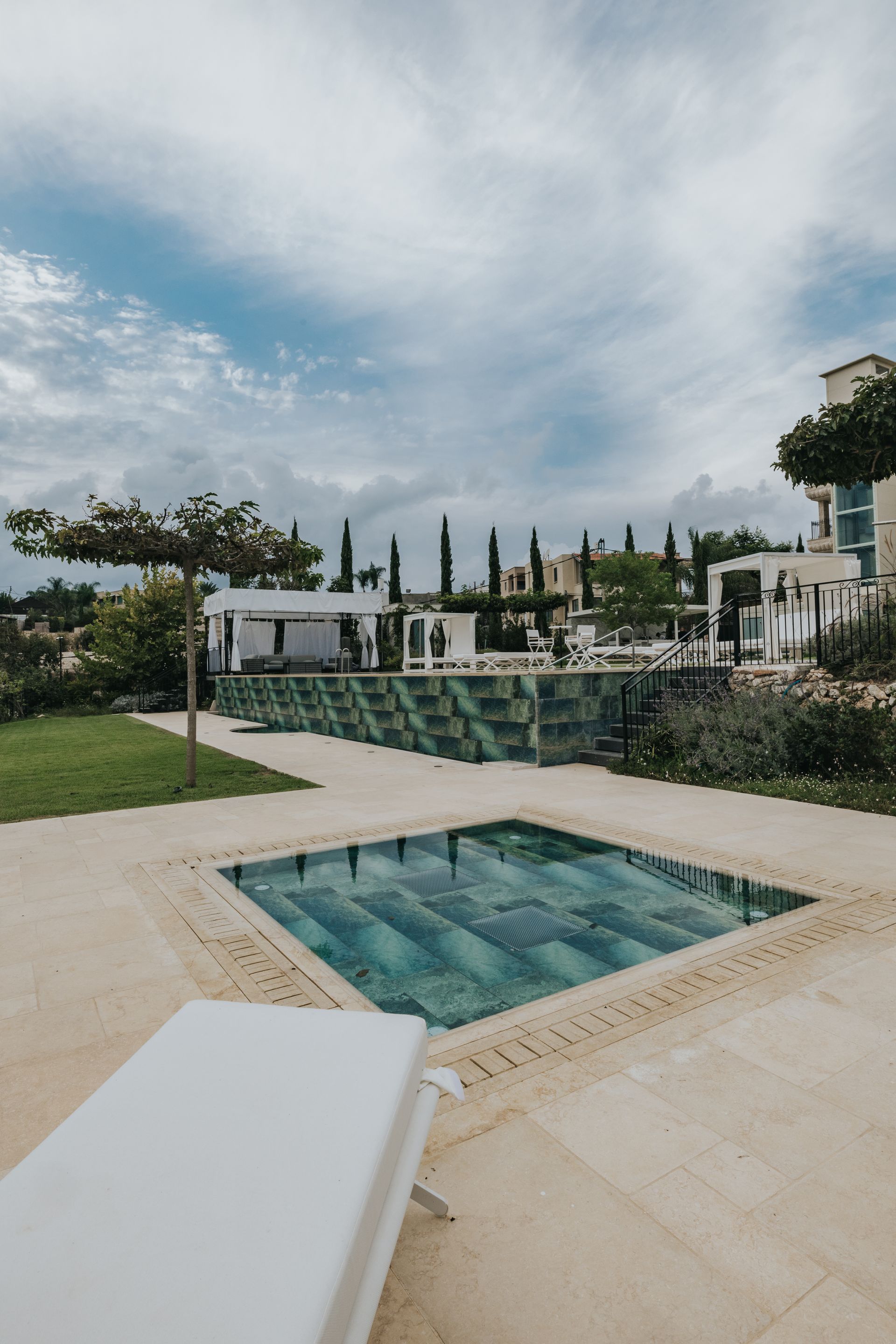 Pool area with green-tiled pools, lounge chair, white buildings, trees, and cloudy blue sky.