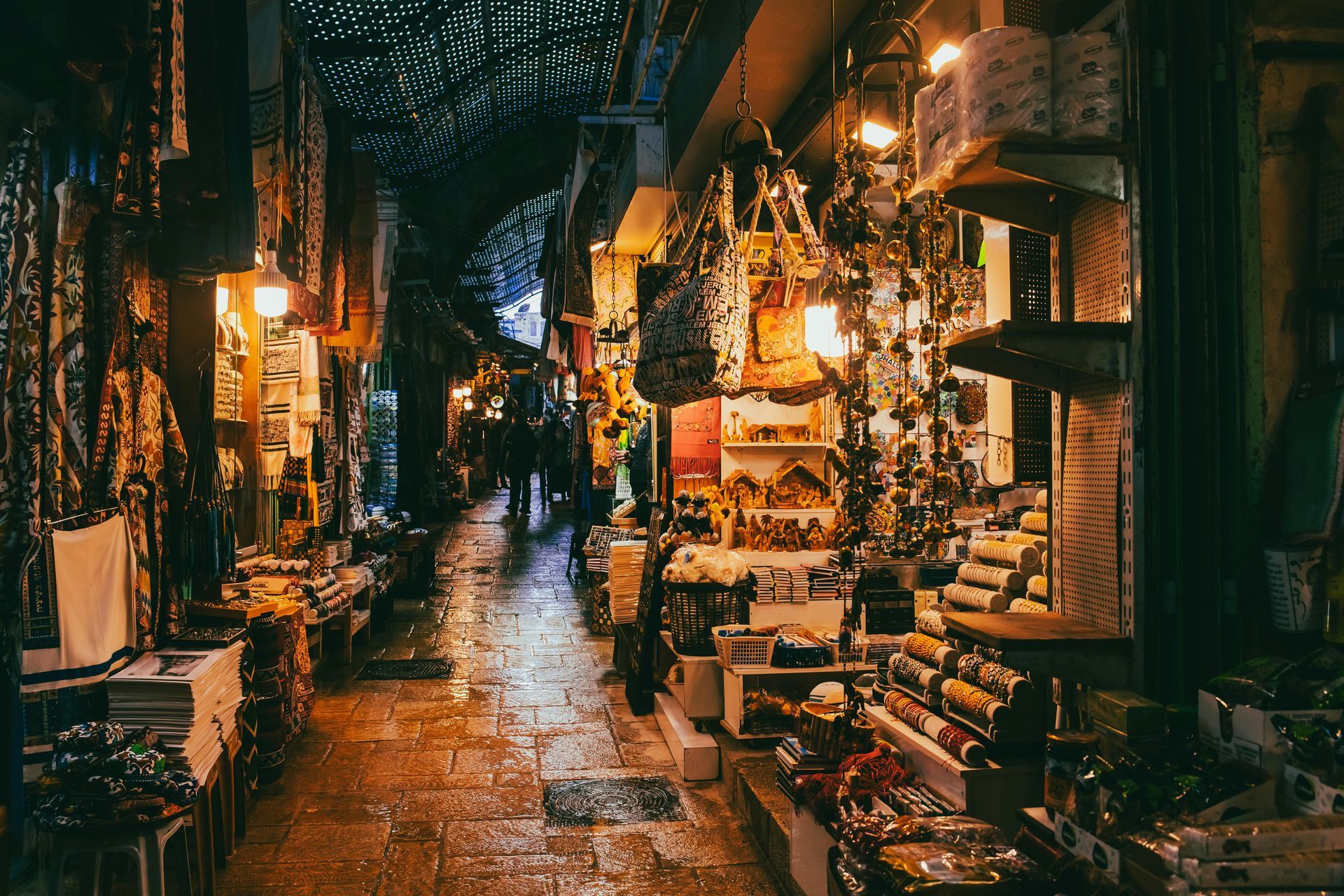 Alleyway market at night; shops with goods line the stone path, illuminated by hanging lanterns.