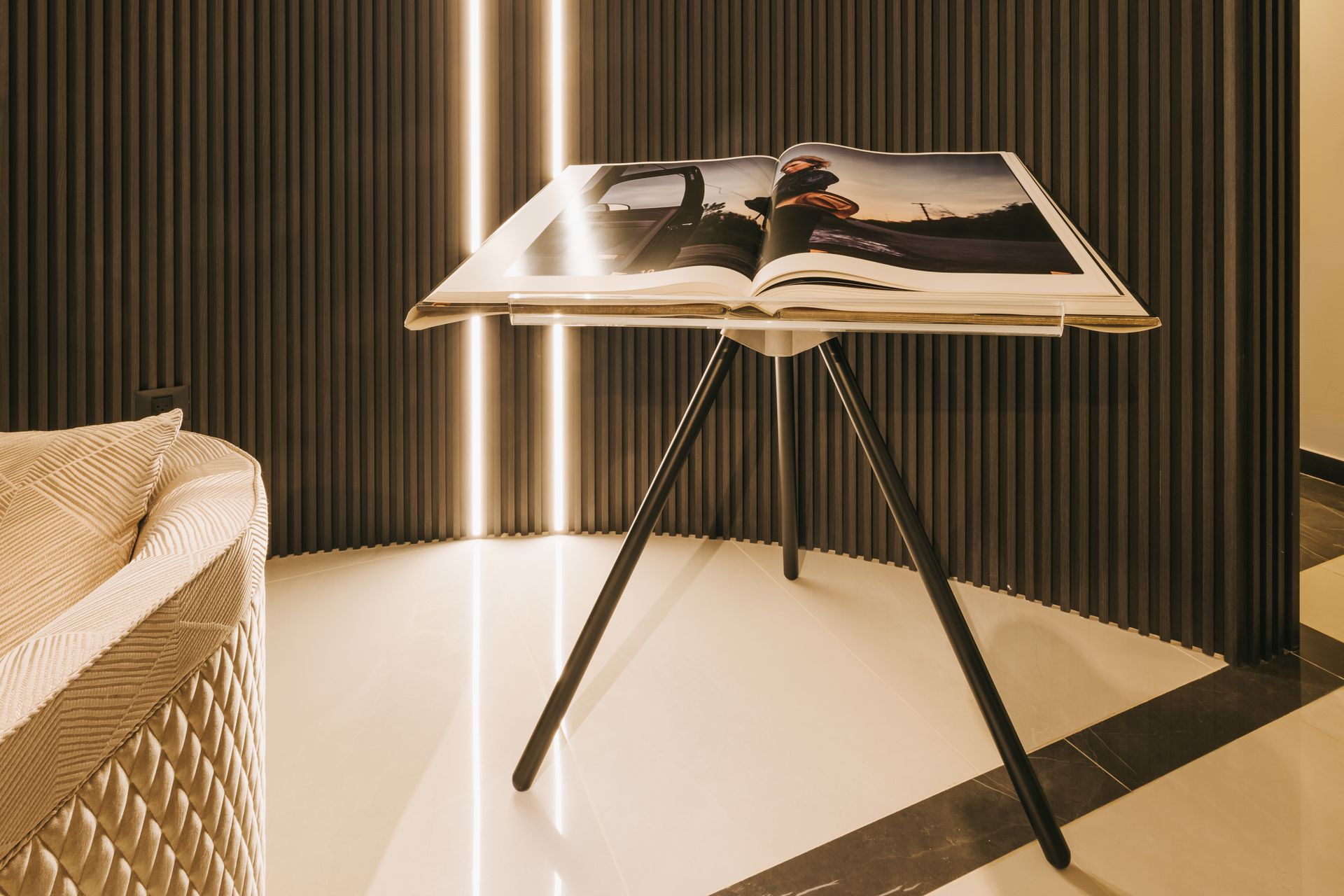 Book on tripod table, against textured dark wall with vertical lights, beige floor, part of sofa on left.