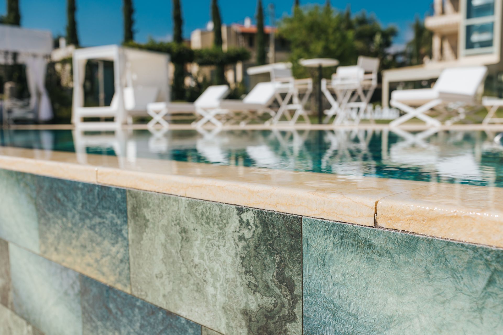 Pool edge with stone tile, blue water, white lounge chairs in the background.