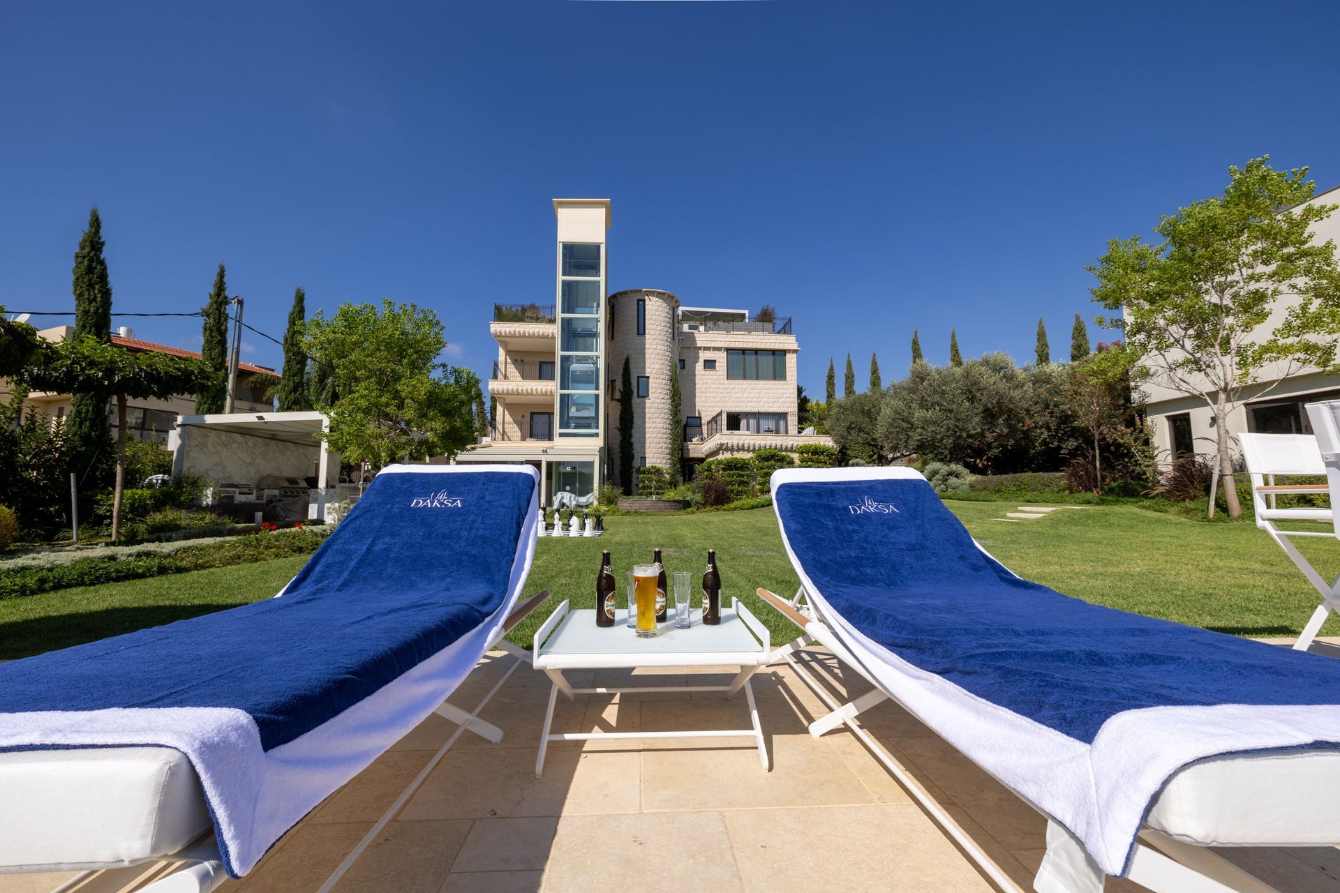 Two blue lounge chairs face a house with a table of drinks on the grass, sunny day.