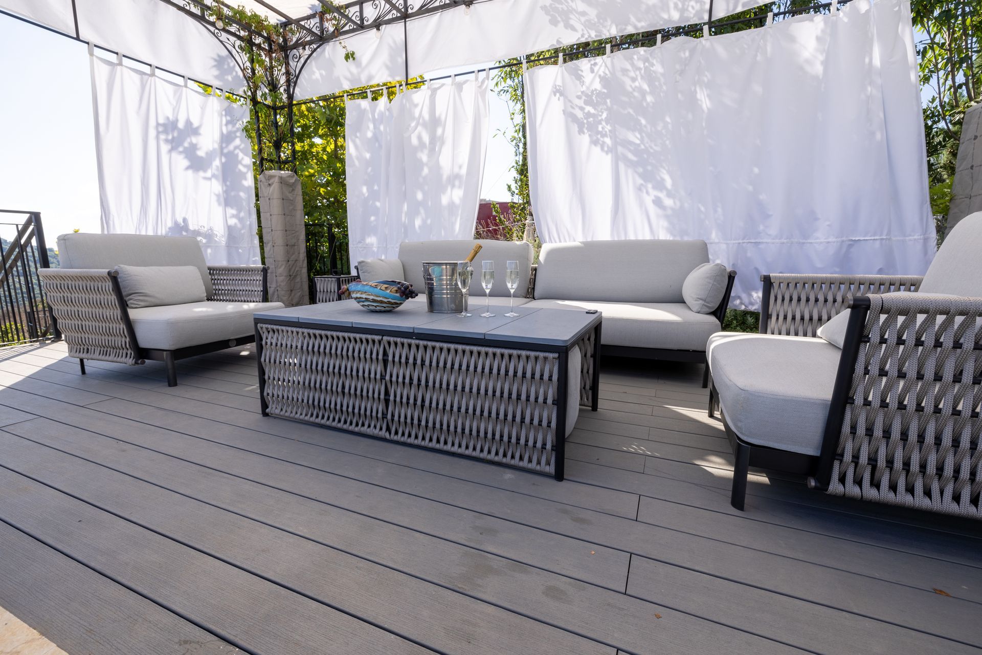 Outdoor seating area with gray furniture on a wood deck, draped with white curtains under a pergola.