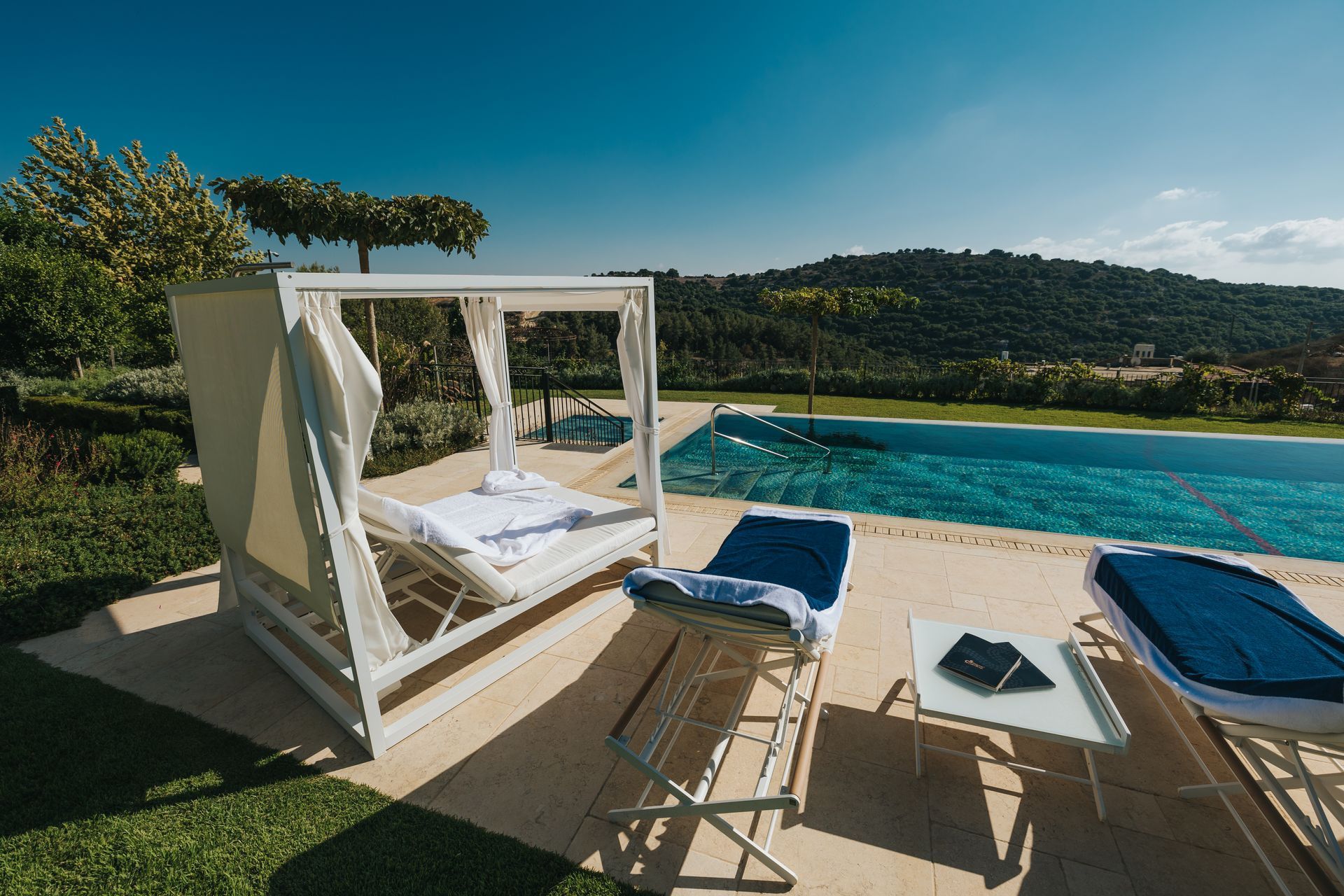 A poolside scene with a white canopy bed, lounge chairs, and a mountain backdrop under a bright sky.