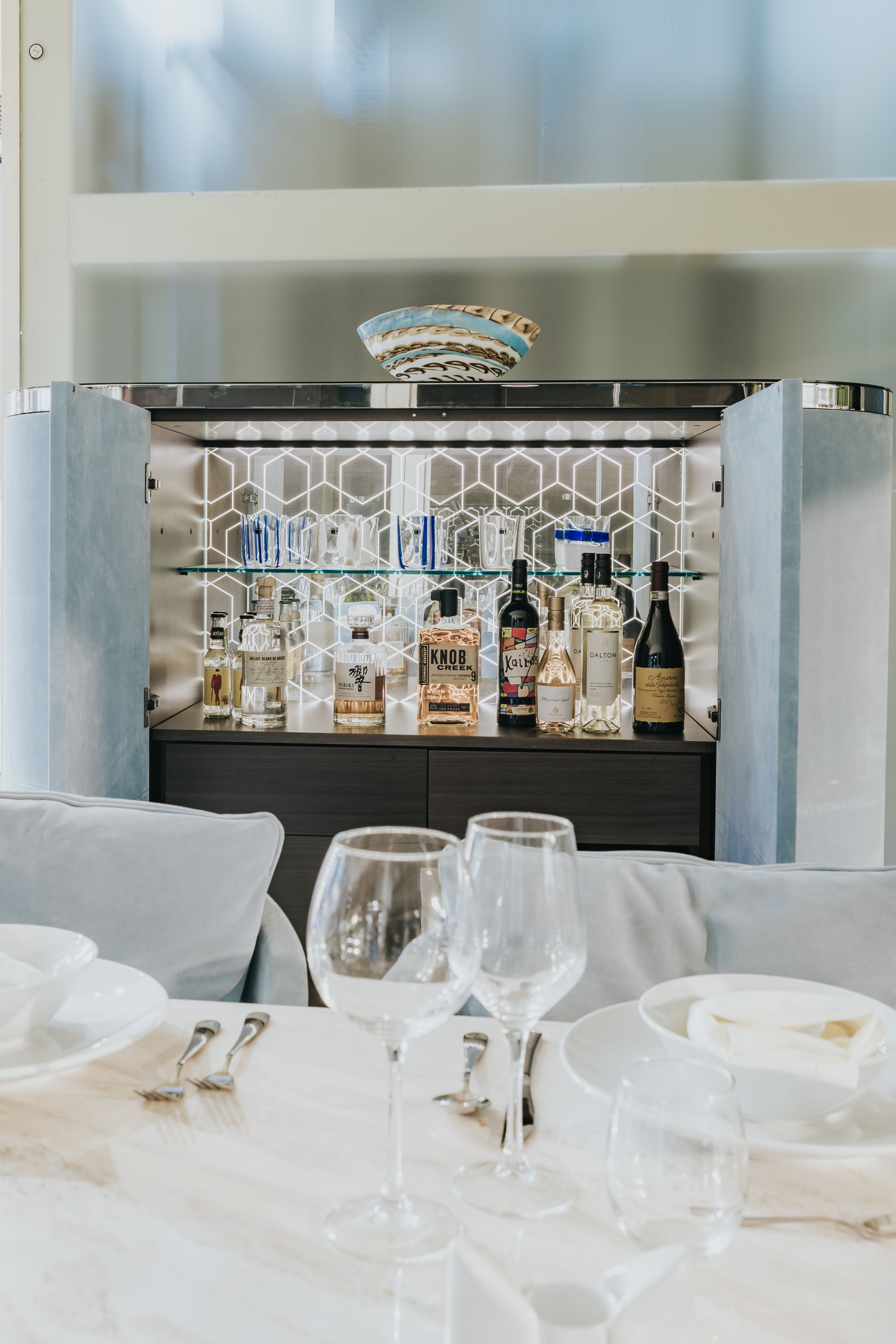 Elegant dining setting with bar. White table, glassware, bottles, and decorative bowl on a dark cabinet with mirrored back.