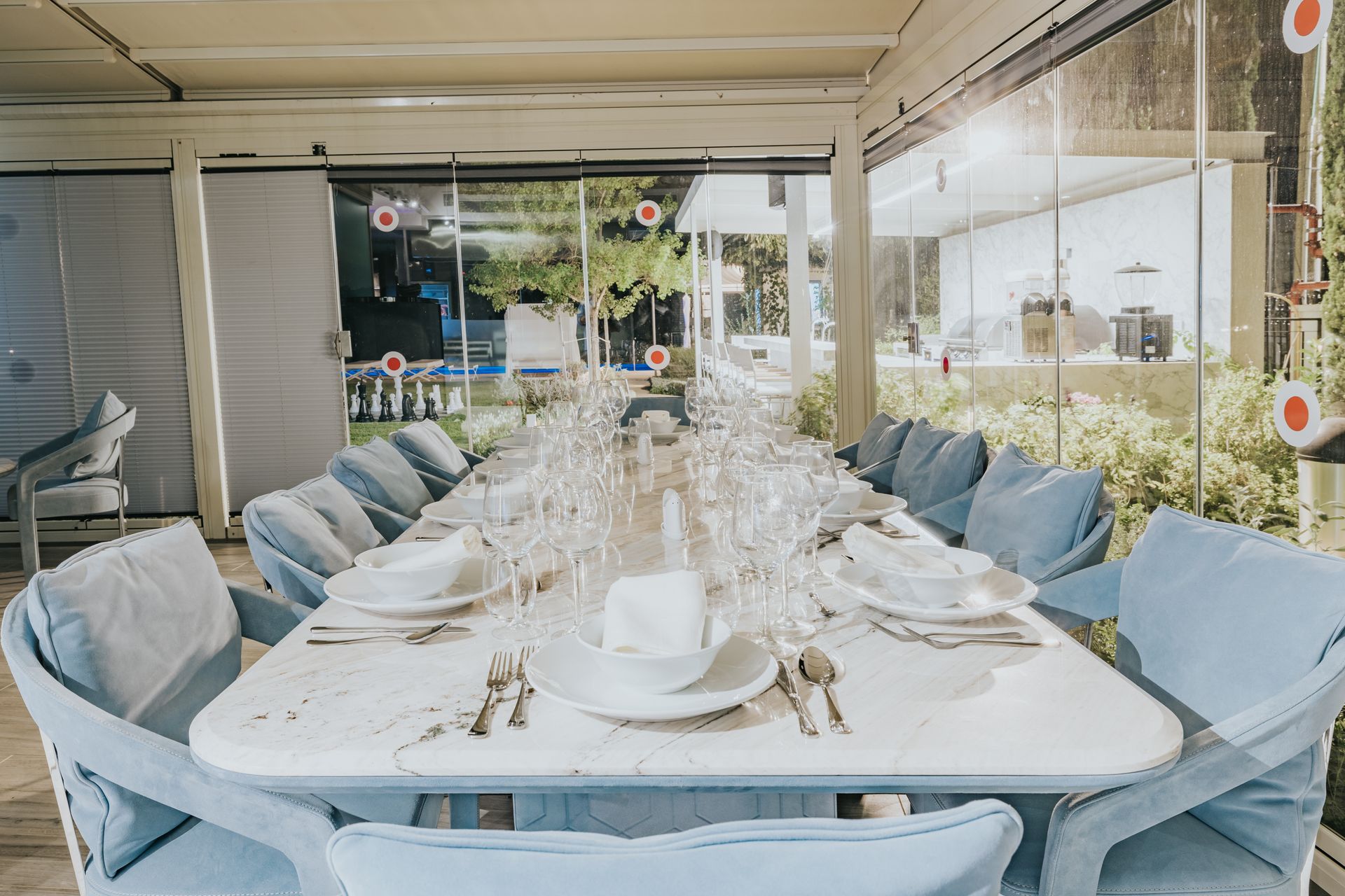 Long dining table set for a meal, with light blue chairs and glassware. Outdoors, clear glass walls.