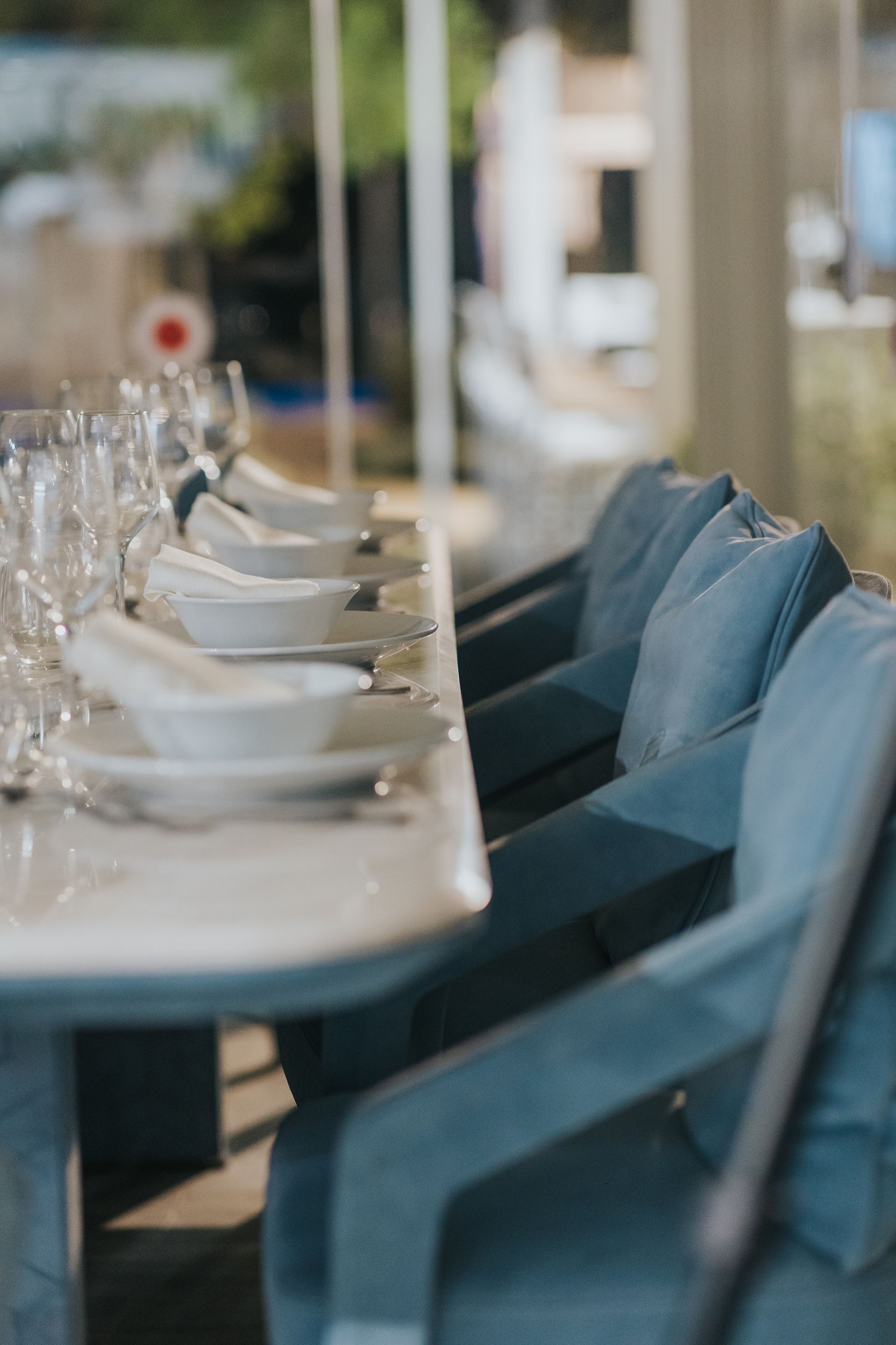 Long table set with white dishes, glasses, and blue upholstered chairs, outdoors.