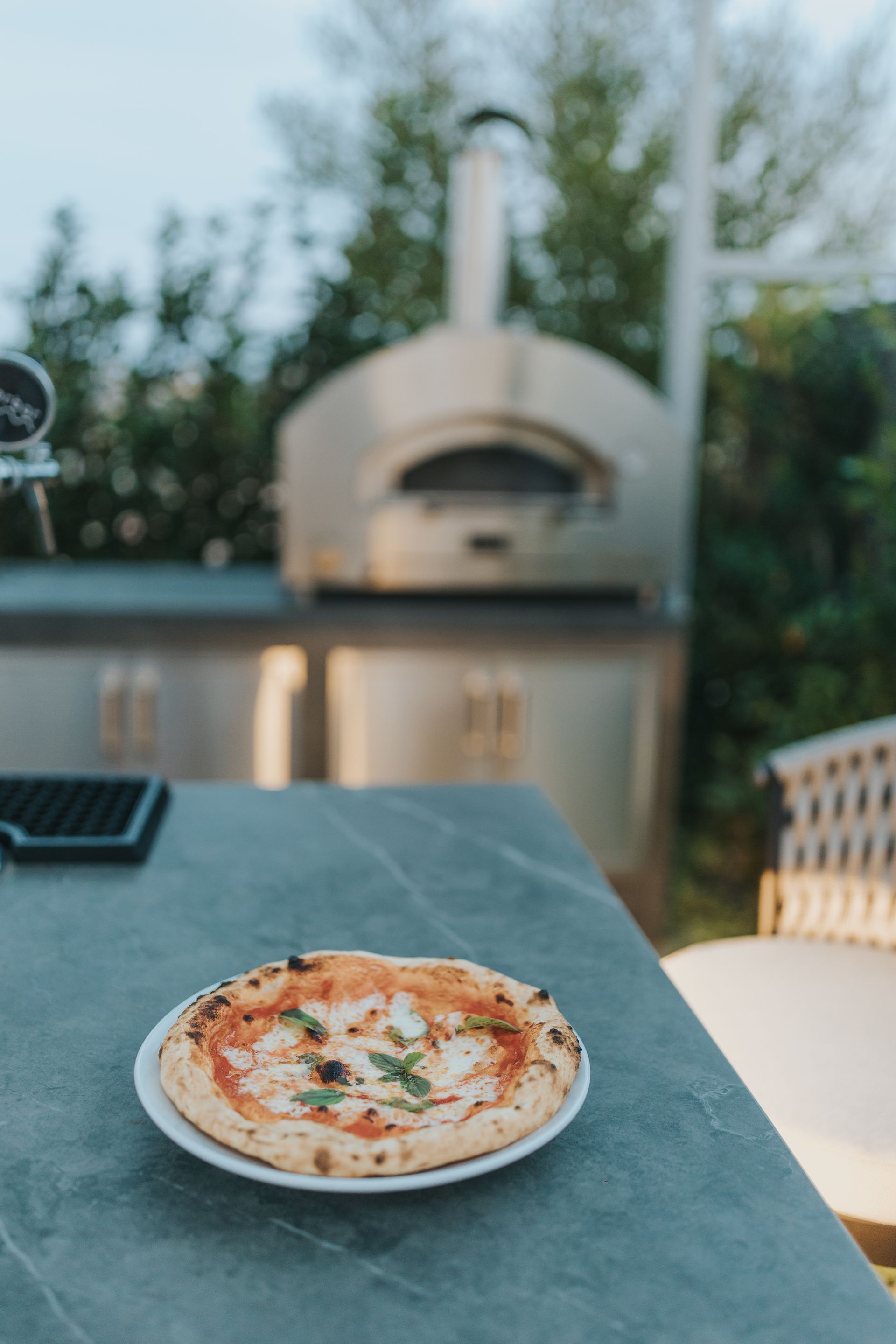Pizza on a plate atop a stone countertop with a pizza oven in the background, outdoor setting.