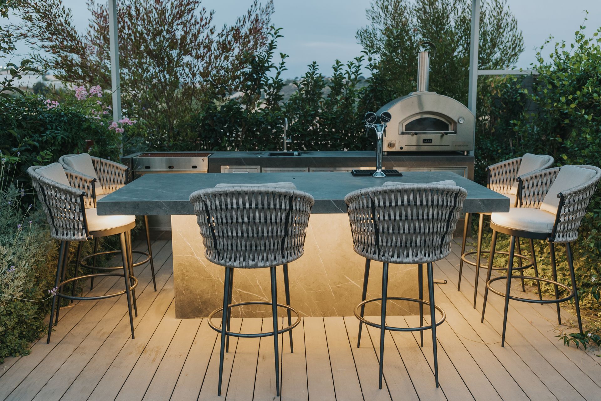 Outdoor kitchen with a pizza oven, bar seating, and lush greenery in twilight.