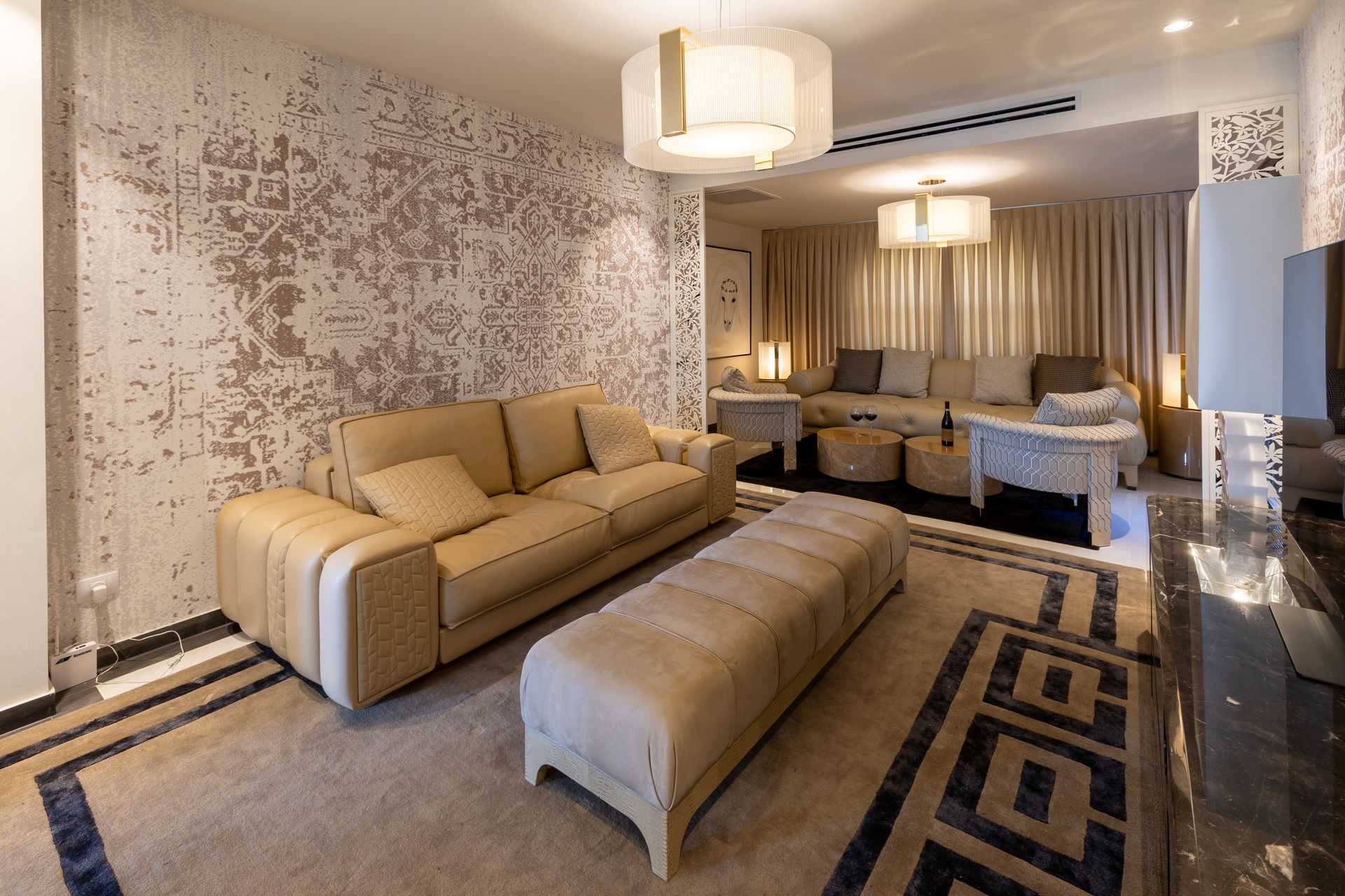 Beige living room with a sofa, ottoman, patterned rug, and decorative wall.