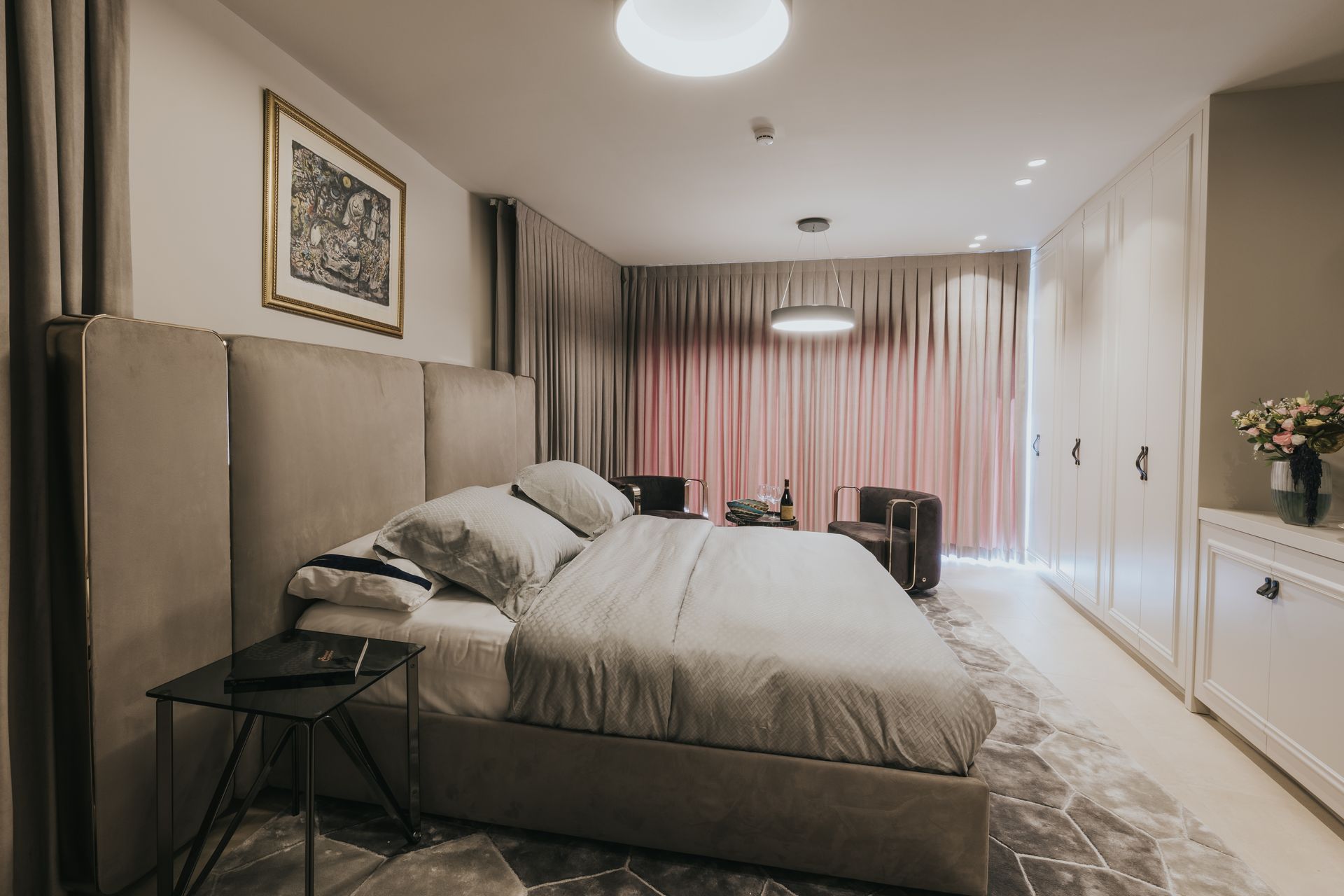 Bedroom with large bed, gray and pink curtains, artwork, and white storage cabinets.