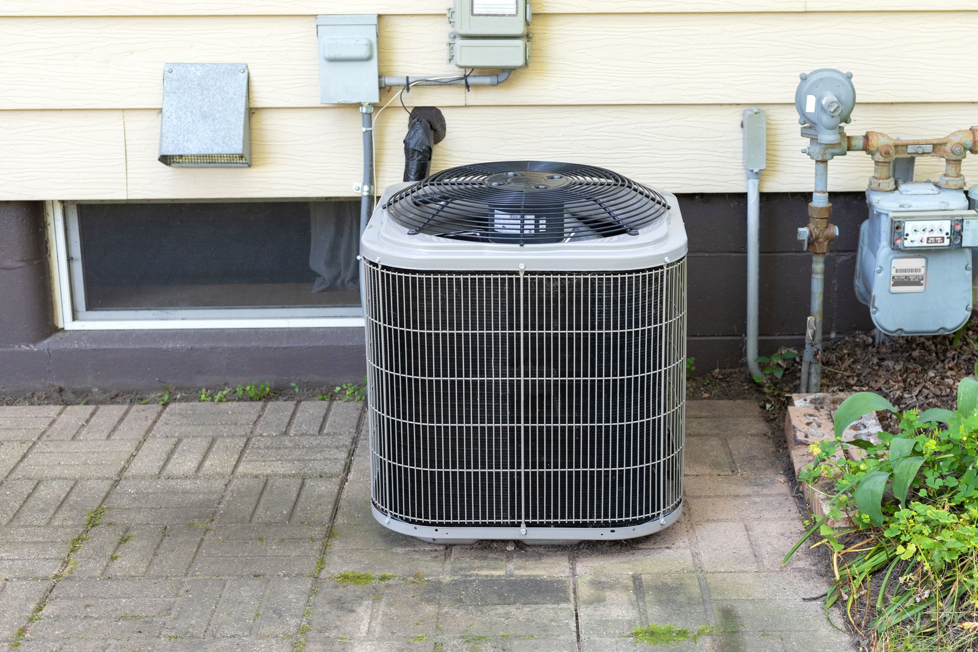 Outdoor air conditioner unit beside a house window, with utility meters and a brick walkway nearby Outdoor air conditioner unit beside a house window, with utility meters and a brick walkway nearby