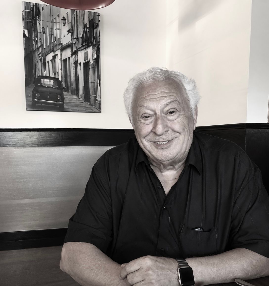 Smiling man seated at a table in a restaurant, wearing a dark shirt with a black-and-white street photo behind him Smiling man seated at a table in a restaurant, wearing a dark shirt with a black-and-white street photo behind him