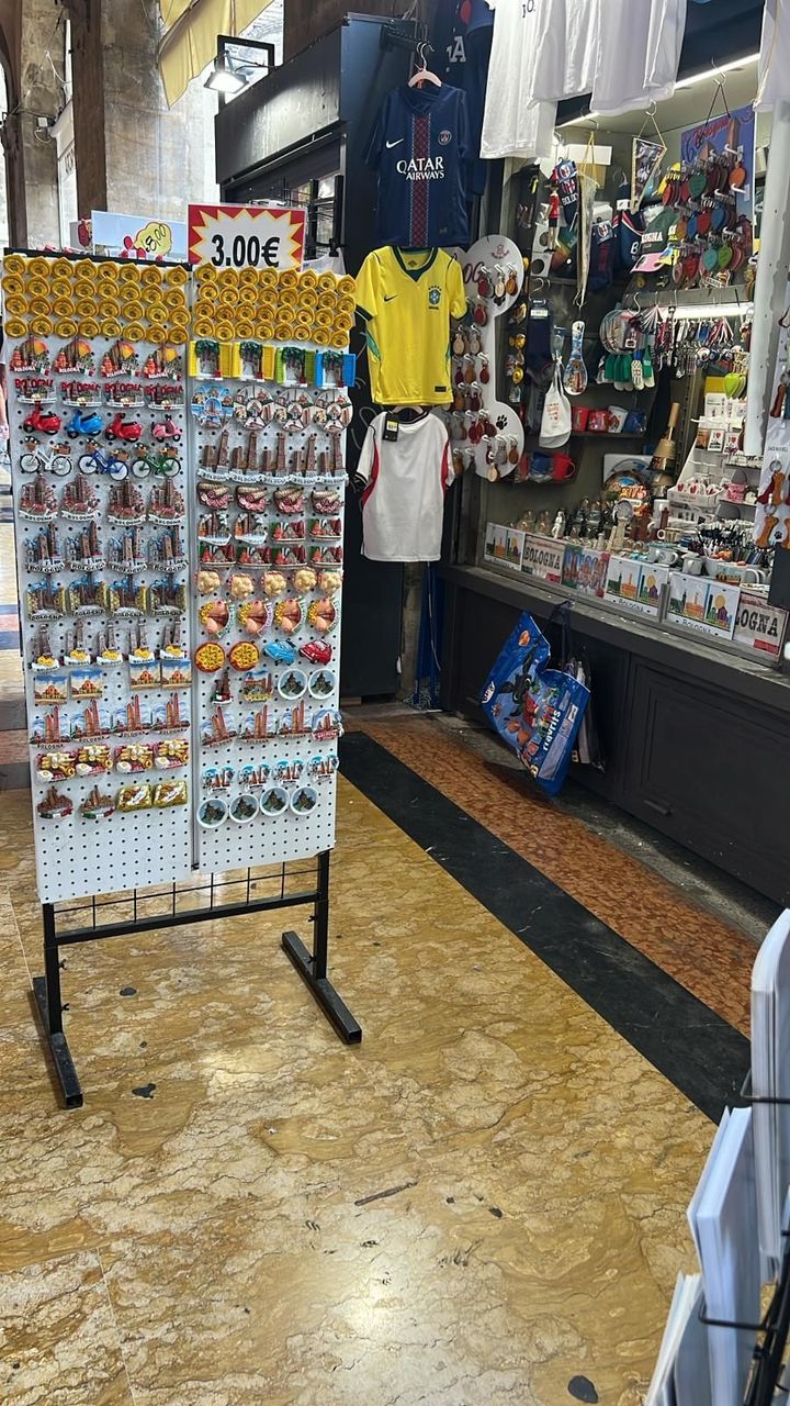 Corridoio del negozio di souvenir con magliette appese, cartoline e un espositore su un pavimento di marmo.