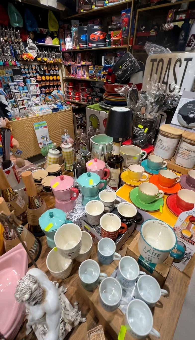 Tazze e utensili da cucina in ceramica colorata esposti su un tavolo affollato in un negozio, con scaffali di merci alle spalle.