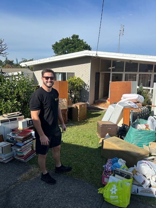 Man standing in a cluttered backyard beside piles of boxes, bags, and household items