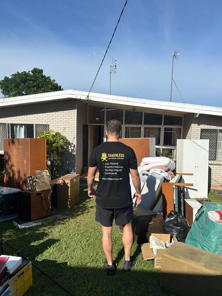 Person carrying items in a grassy yard beside a single-story house with boxes and furniture outside.