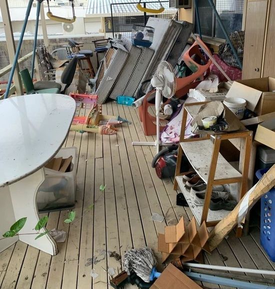 Cluttered porch with broken furniture, boxes, and debris on a wooden floor