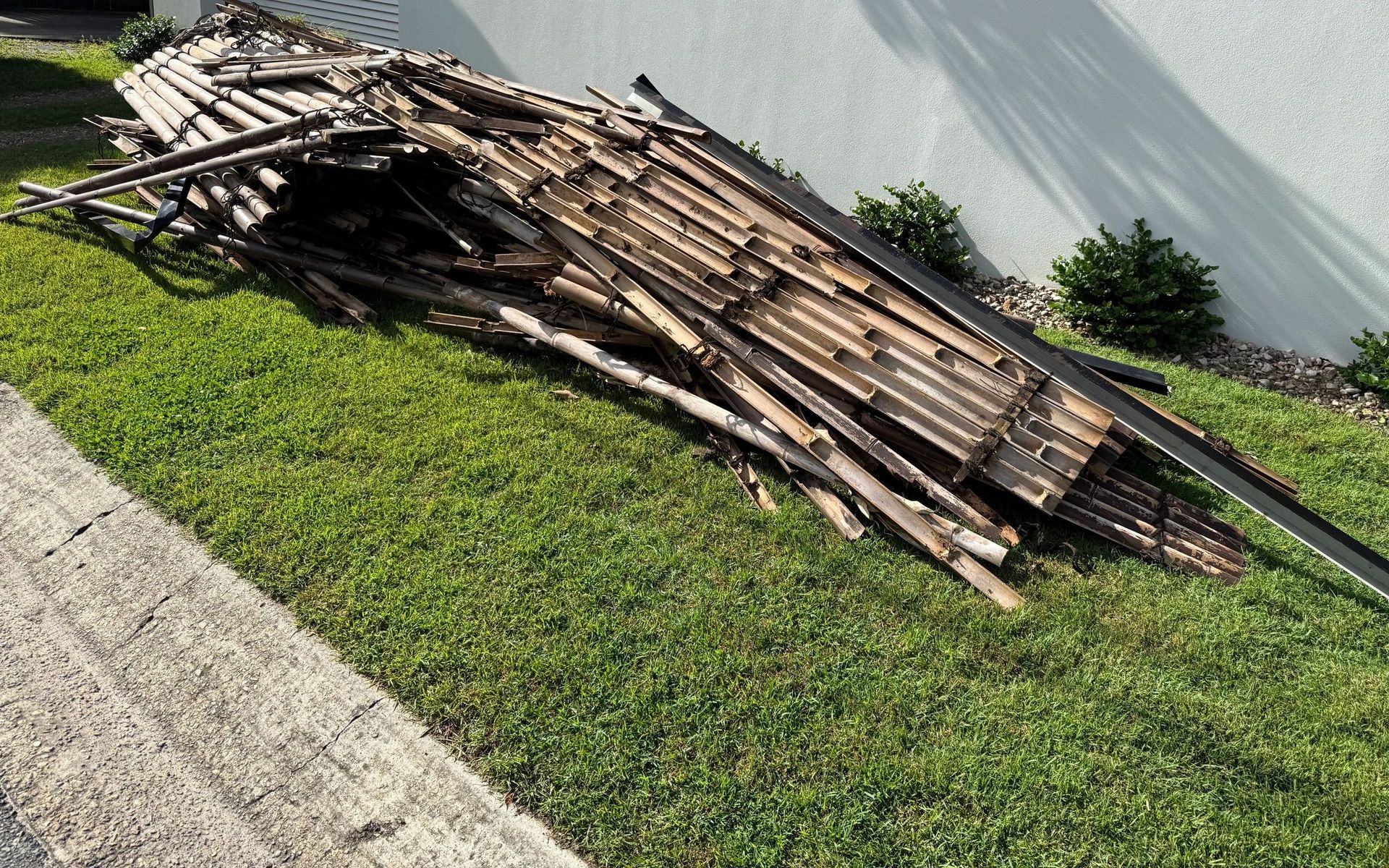 Pile of broken wooden planks on a grassy verge beside a curb and wall