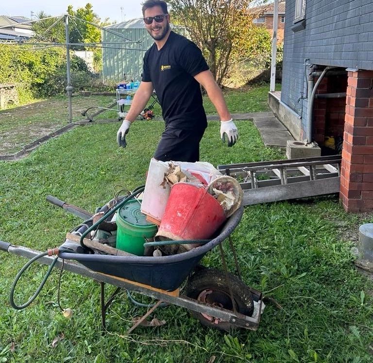 Man pushing a wheelbarrow full of yard tools across a grassy backyard near a house and ladder