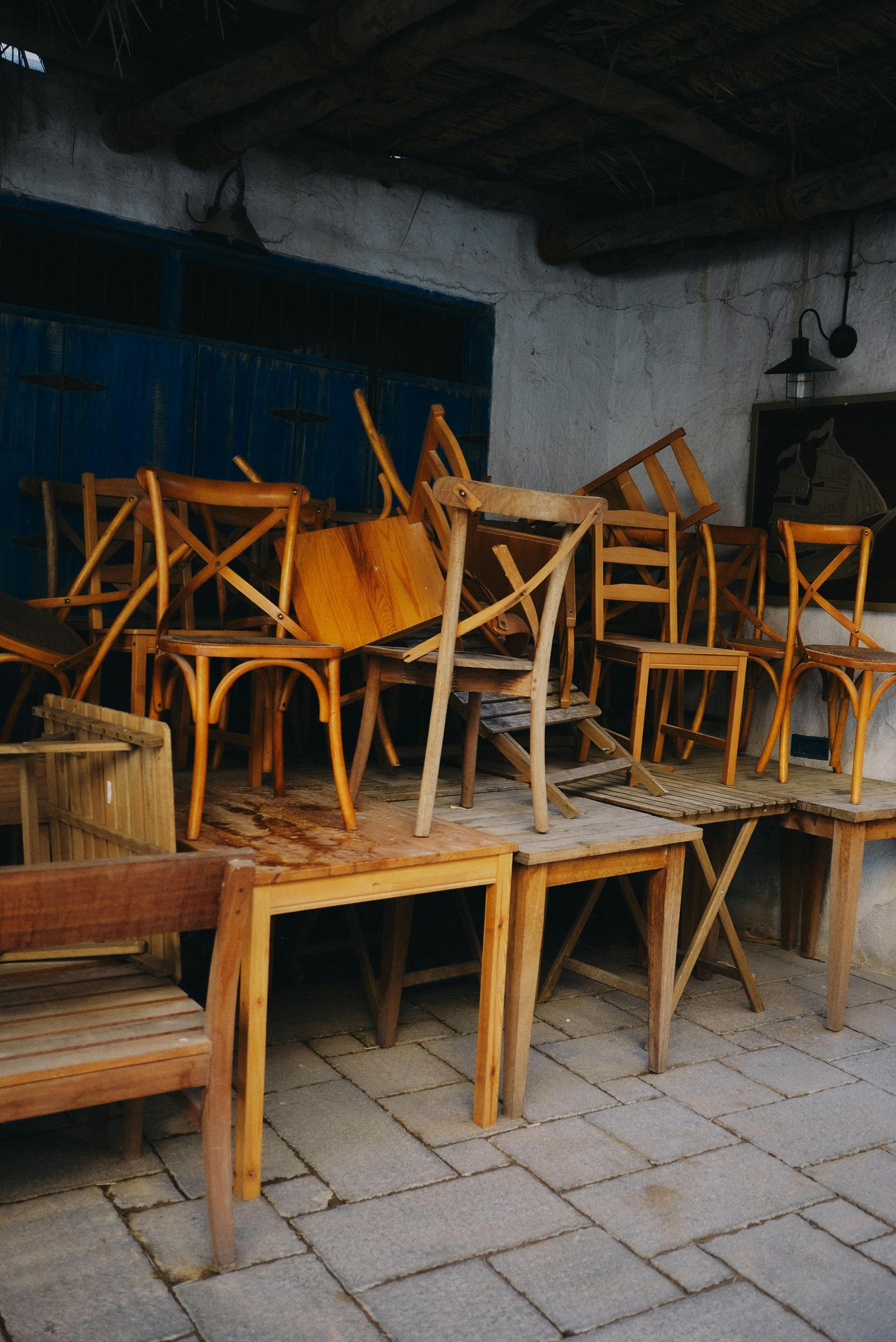 Stacked wooden chairs and benches in a dim indoor storage area with tiled floor.