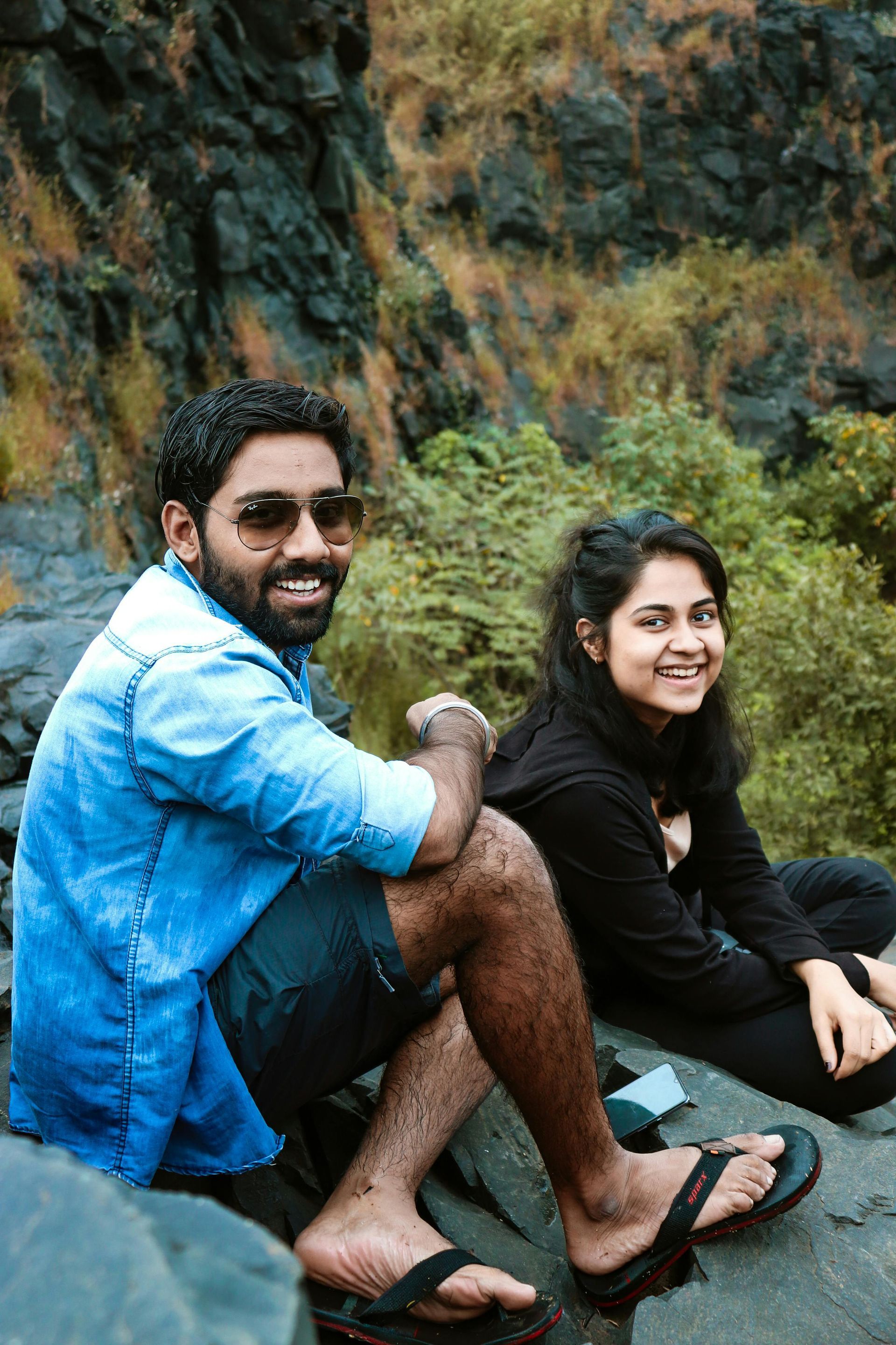 Two people sitting on a rocky ledge outdoors, smiling at the camera.