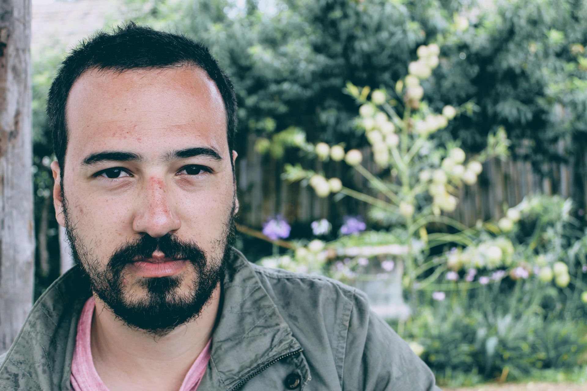 Man with beard in a green jacket standing outdoors with blurred greenery behind him