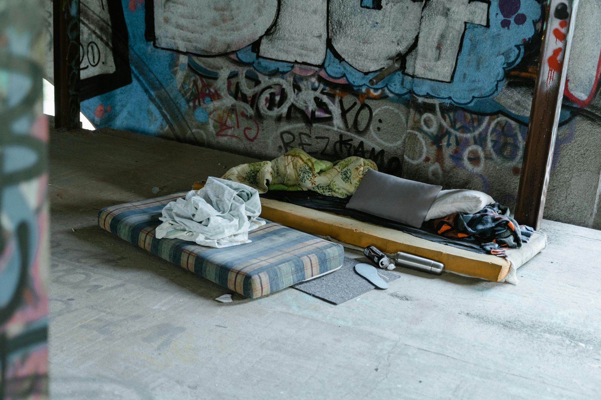 A mattress with folded clothes and a long wooden object on a concrete floor in a graffiti-covered room.