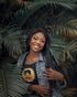 Smiling woman in denim jacket standing among large palm leaves, holding a strap on her shoulder.