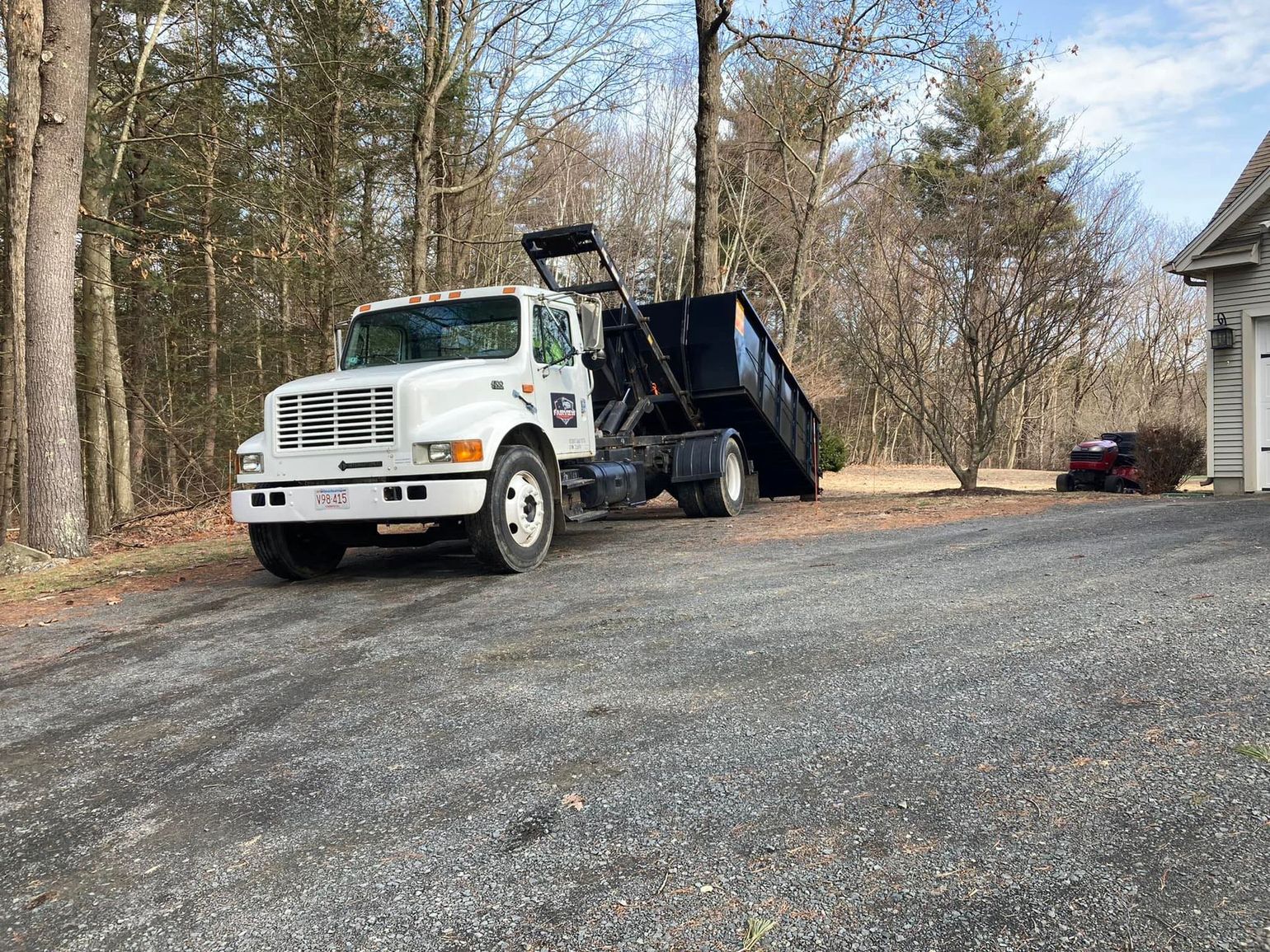 White dump truck with black container on a gravel driveway in front of a house.