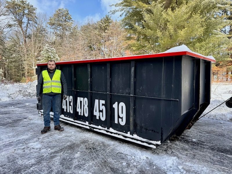 Man in a yellow vest stands beside a large black dumpster in a snowy outdoor setting. 
