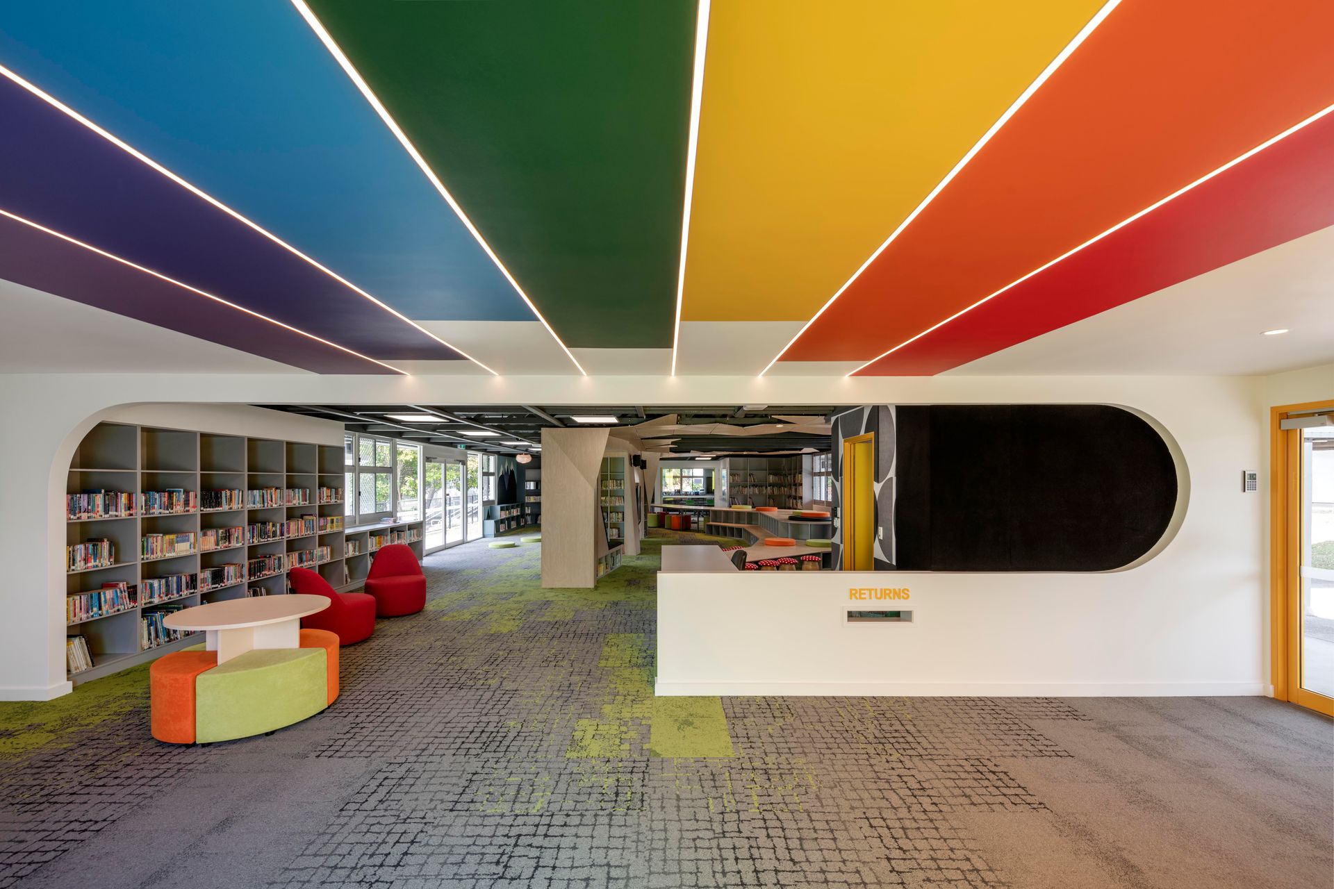Library interior with rainbow-colored ceiling stripes and built-in bookshelves. A reception desk and seating are visible.