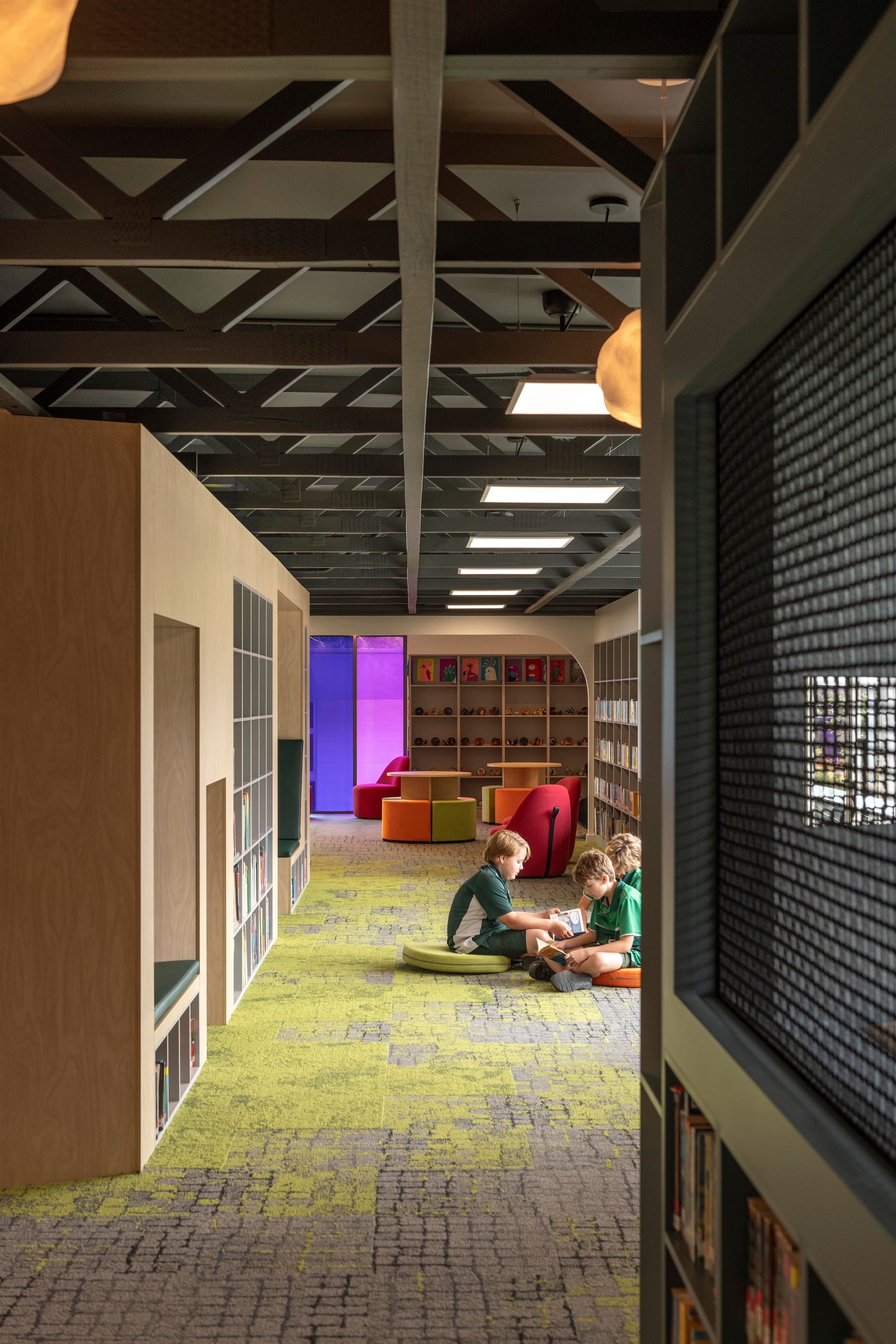 Children sitting on mats in a library with books. Light green carpet, colorful seating, and wooden beams.
