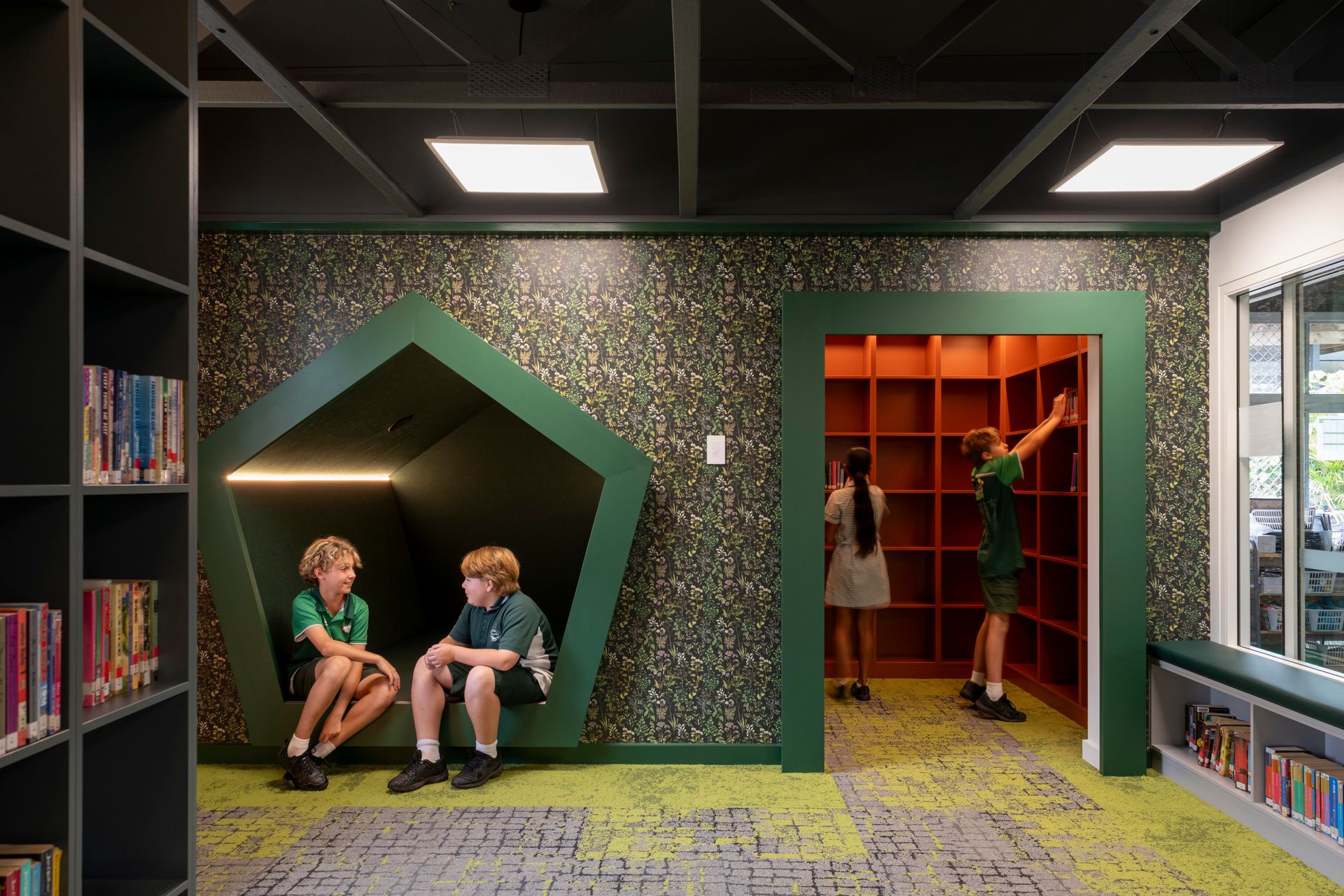 Children in a school library with book shelves. Green accent walls, one with a reading nook, a square bookshelf, and children reaching for books.