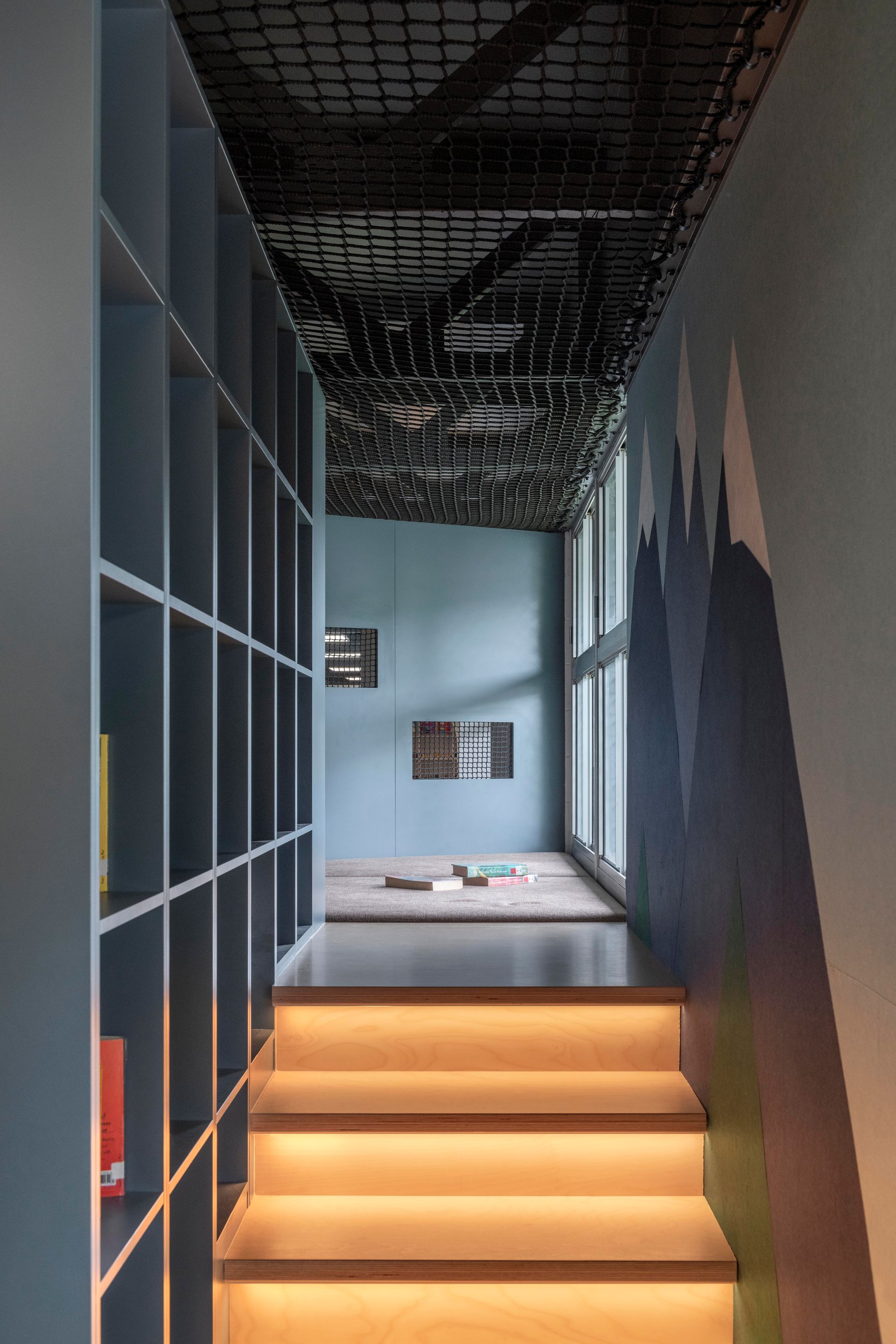 Stairway with warm lighting leading to a blue room with bookshelves and mountain mural on the wall.