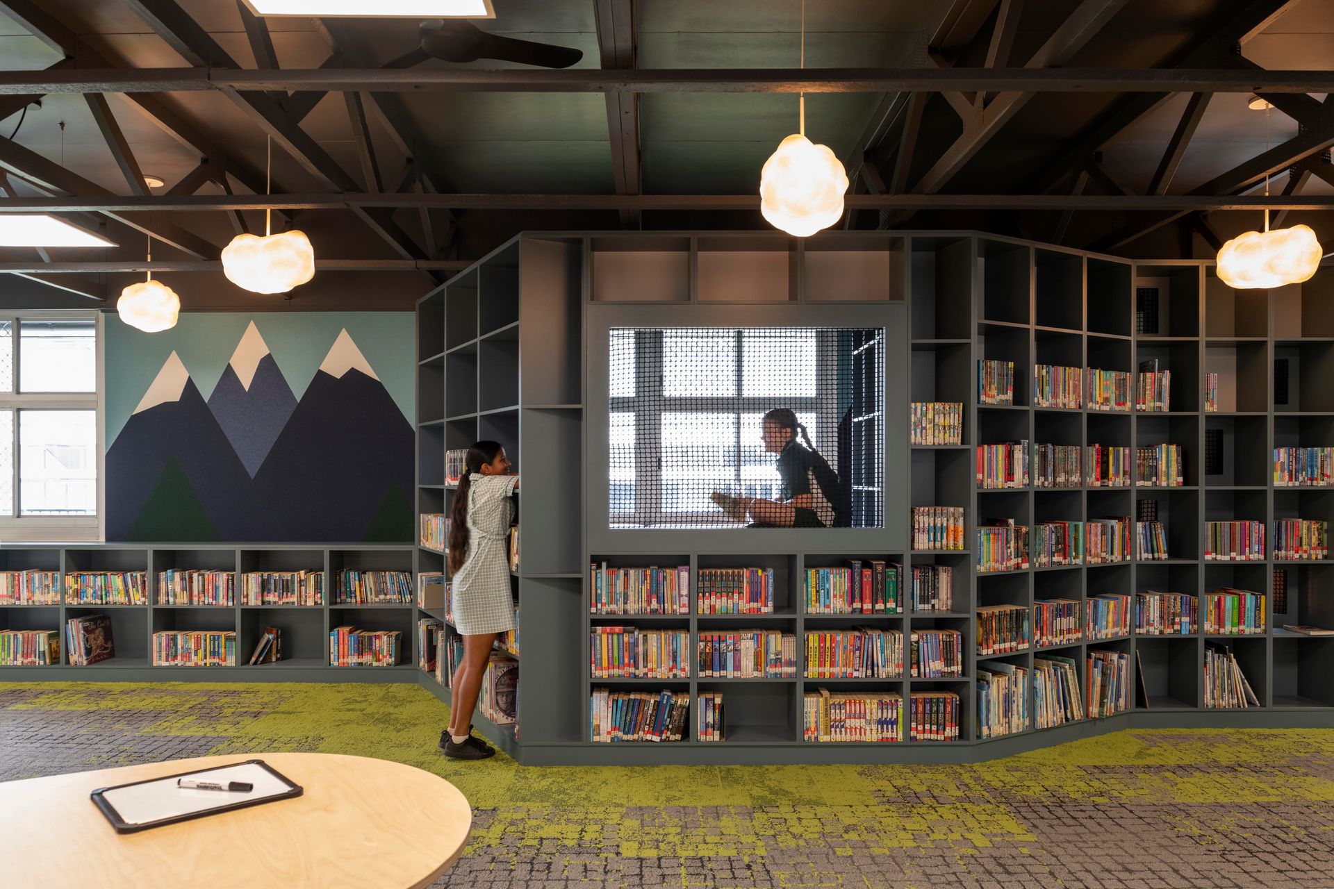 Library interior with bookshelves, reading nook, and two people. Green carpet, mountain mural, and hanging lights.