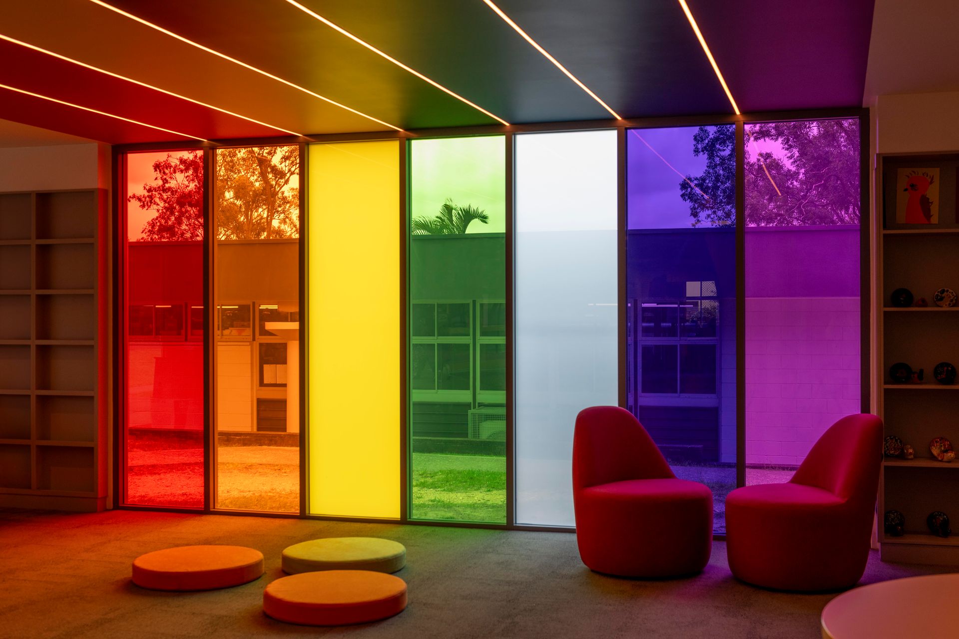 Room with rainbow-colored window panels; two red armchairs and round floor cushions.