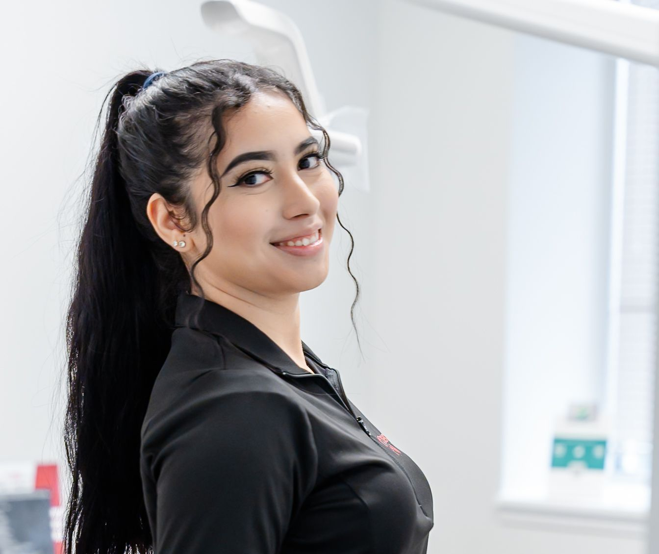 A person with dark hair in a ponytail smiles, looking over their shoulder in a bright, professional office setting.