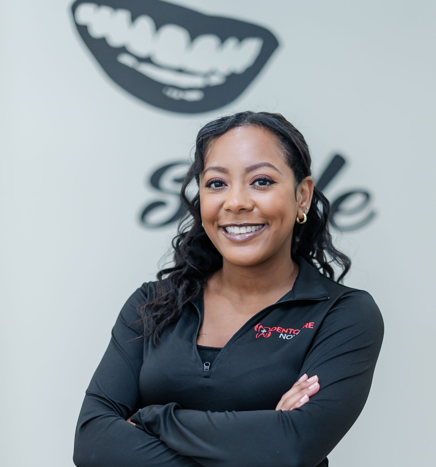 A smiling person with long dark wavy hair stands with arms crossed, wearing a black zip-up top against a branded wall.